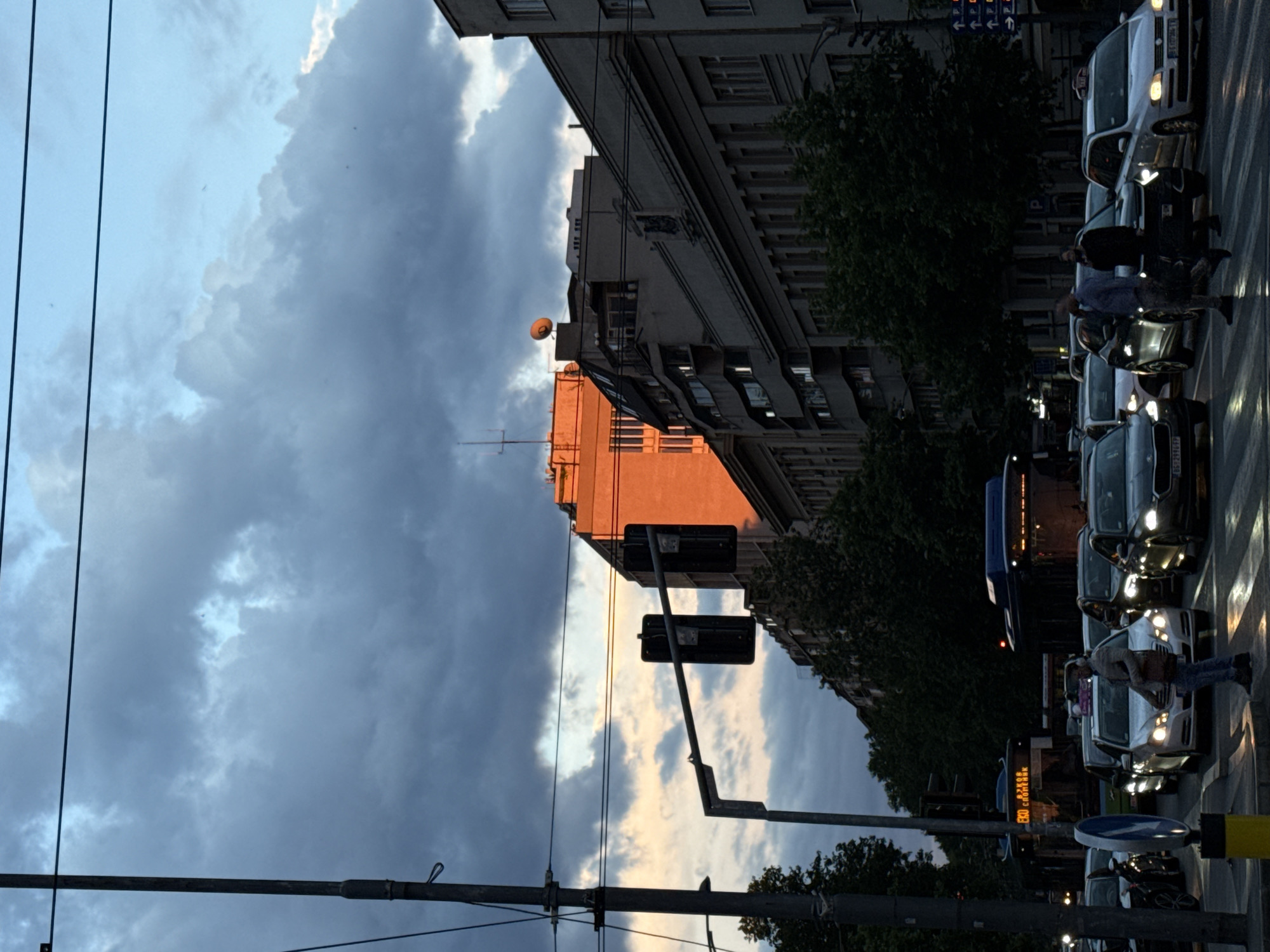 The image depicts a city street scene in Belgrade, Serbia, during what appears to be dusk. The sky is a mix of deep blues and grays from looming storm clouds, with patches of warm, orange-tinged light from the setting sun illuminating some of the buildings. The architecture suggests a European city with multi-story buildings lining the street.
In the foreground, there is a busy intersection with a traffic light. Several cars are stopped at the intersection, suggesting traffic congestion. A person is crossing the street in the foreground, wearing headphones and carrying a bag. Another two men are also visible near the crosswalk, appearing to be walking or standing.
A bus with illuminated signage is stopped in the background, indicating public transportation is active. Utility poles and overhead wires are visible, characteristic of urban infrastructure. The overall atmosphere is somewhat dramatic due to the lighting and cloud cover, but the scene is primarily a snapshot of everyday urban life.
