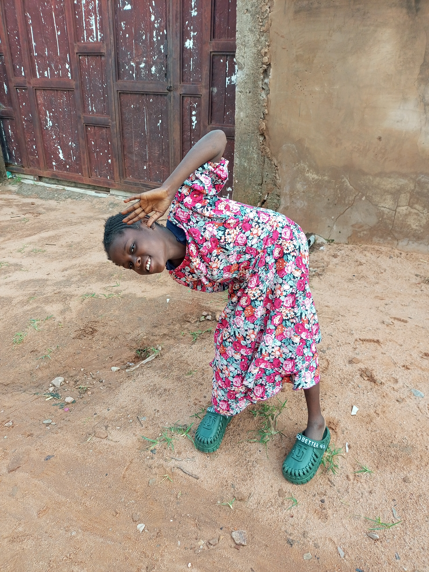 A young Black girl, wearing a floral-print dress and green clogs, bends over with her head down and right arm raised, her left hand near her ear. Her mouth is open in a smile, and she looks towards the viewer. She is in an outdoor setting with a sandy ground that has sparse green grass and some scattered pebbles and debris. Behind her is a rough, weathered concrete wall on the right and a large, dark brown wooden door with vertical planks and horizontal crossbars on the left. The overall scene appears to be in a rustic or underdeveloped area.
