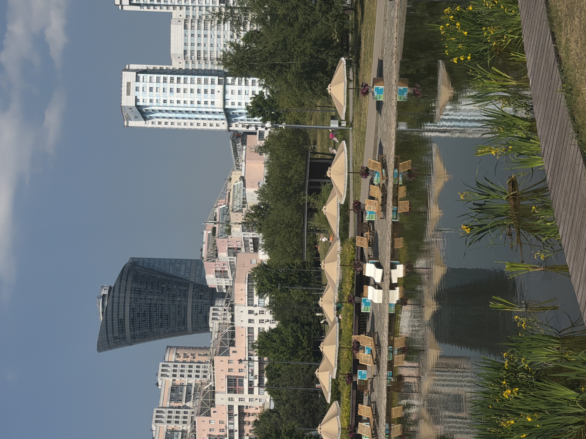 In Khimki, Russia, a city park scene unfolds with a pond in the foreground, featuring tall green reeds and yellow flowers. The pond's surface reflects the surrounding environment, including the sky, buildings, and trees. Along the pond's edge, a wooden walkway leads the eye towards the background. Further back, a paved area is lined with rows of empty sun loungers and scattered beige umbrellas, indicating a recreational space. Beyond this, a line of lush green trees forms a backdrop, separating the park amenities from the urban landscape. In the distance, a cluster of modern buildings stands out against the clear blue sky. The most prominent of these is a unique, curved, dark glass skyscraper, a distinctive architectural feature of the city. Other taller residential buildings with light-colored facades and repetitive window patterns are also visible. The overall scene suggests a sunny day in an urban park with amenities for relaxation and leisure, set against a backdrop of contemporary city architecture.