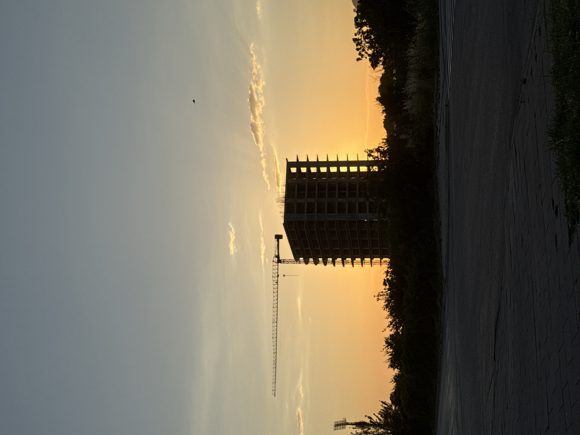 The image depicts a tall, unfinished concrete building under construction in Bar, Montenegro. A crane is attached to the building, silhouetted against an orange and yellow sunset. The sky ranges from light blue to orange. There are a few small clouds in the sky. Vegetation is visible along the right side of the image. A dark asphalt surface is visible in the lower right corner. There is a dark object, possibly a bird, in the sky.