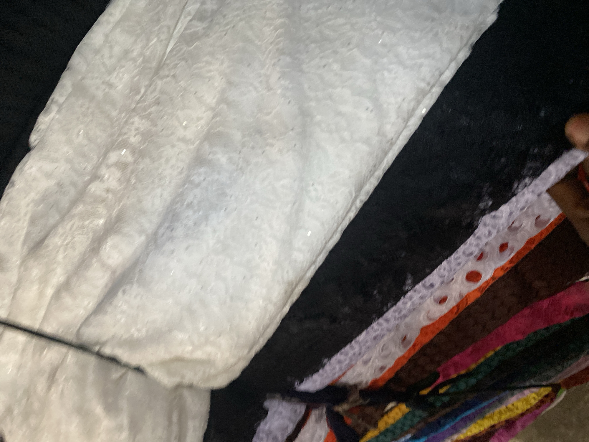 This image depicts a close-up view of stacked textiles in Jalingo, Nigeria. The most prominent item is a white, patterned fabric, possibly lace or brocade, with a slightly textured surface that appears to have subtle embellishments, such as small, reflective dots or sequins. It is draped over a black fabric base. To the left and below, a colorful array of fabrics is visible, appearing to be folded or rolled. These fabrics display a variety of colors, including purple, orange, red, yellow, green, and dark blue, suggesting a market or textile shop setting. The lighting in the image is somewhat dim, creating soft shadows and highlighting the textures of the fabrics. The overall impression is one of abundant and diverse textile materials.
