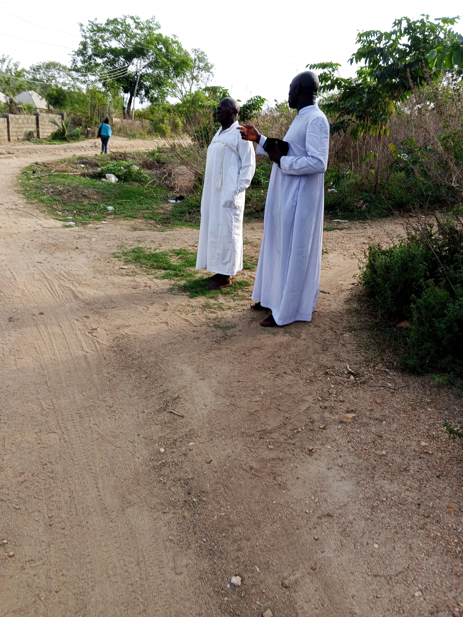 Two dark-skinned men wearing white robes stand on a dirt road. The man on the right holds a black object in his left hand and points with his right. A person wearing a blue shirt walks in the distance on the same dirt road. The location is Zango-ndaji, Nigeria. Vegetation and trees surround the scene.