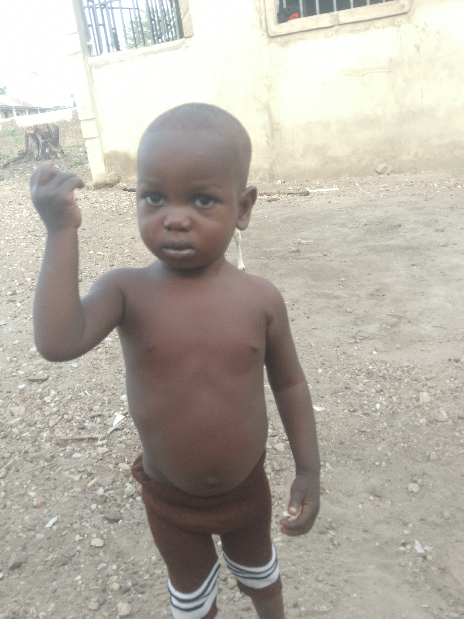 A young, dark-skinned child with a bare chest stands in an outdoor, likely rural, environment. The child has short-cropped hair, and is wearing short brown pants with white-striped socks. The ground is covered in small gravel and dirt. A pale building with a barred window is visible in the background. In the background, there is a stump or tree trunk with some sparse vegetation. The city and specific location are unknown.
