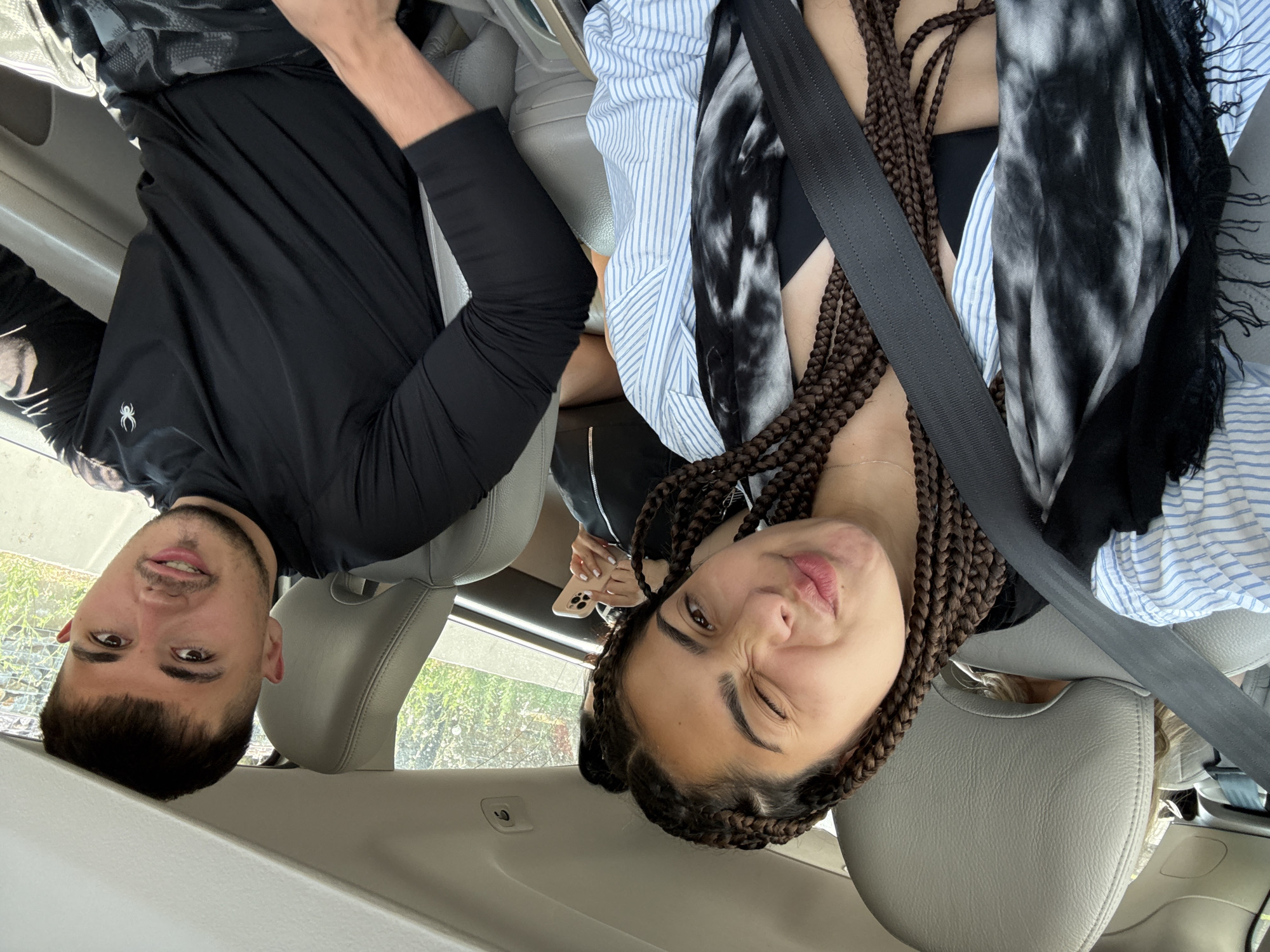 Two individuals, a young woman and a young man, are seated in the front seats of a car. The woman, on the left, is looking towards the camera and making a playful face, winking and pursing her lips. She has dark, braided hair and is wearing a blue and white striped open shirt over a black top, with a black and white patterned scarf. A black seatbelt is visible across her chest. The man, on the right, is looking towards the camera with a slight smile. He has short, dark hair and is wearing a black long-sleeved shirt and camouflage-patterned shorts. The interior of the car, including light gray leather seats and a white headliner, is visible. Outside the car windows, greenery and what appears to be a stone wall are present, suggesting an outdoor setting, possibly in a town or rural area. The image is taken from the perspective of someone in the passenger seat, looking towards the driver and the woman.