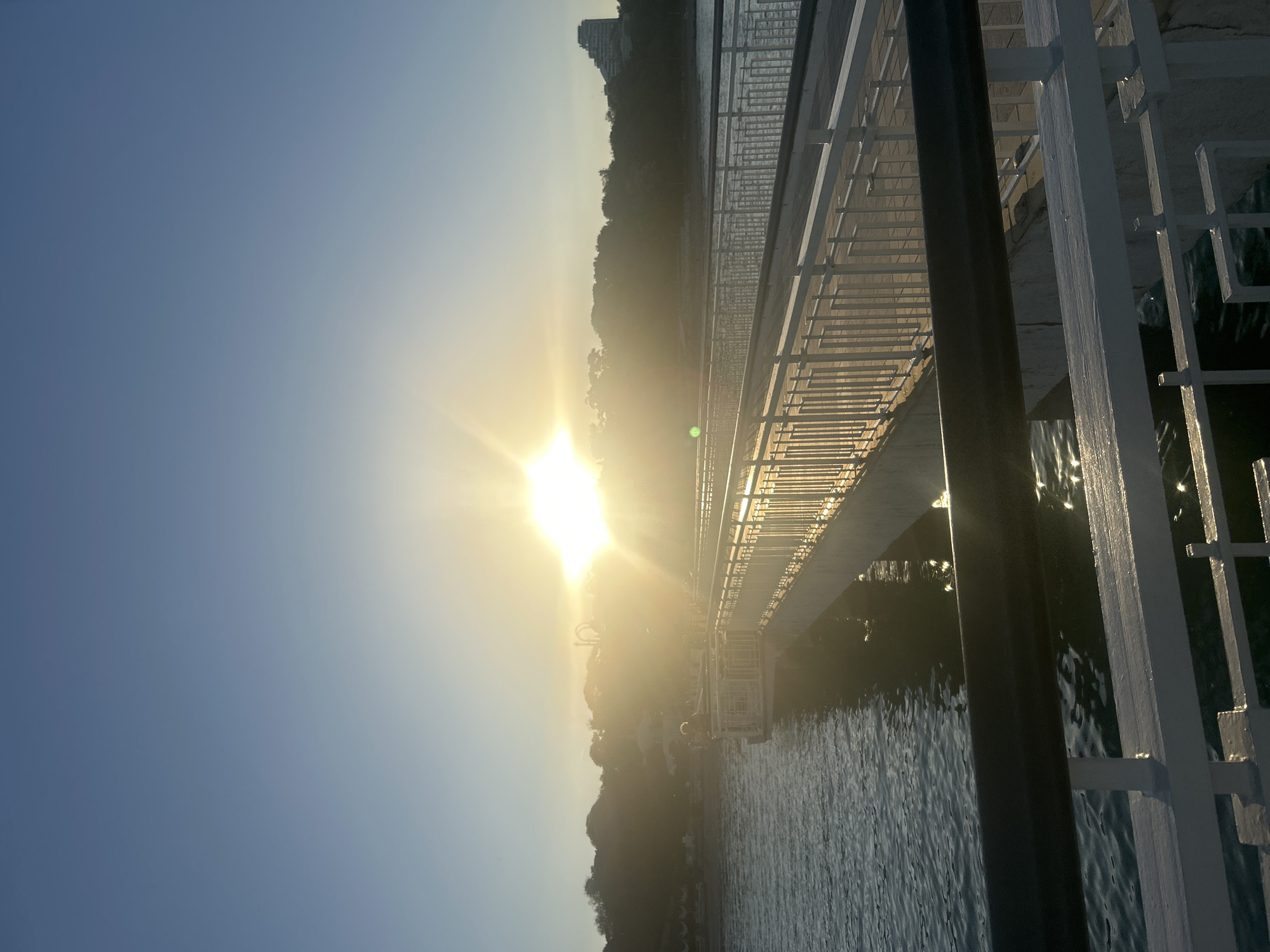The image shows a view of a bridge over water, likely a lake or the sea, at sunset. The sun is visible, partially obscured by the landscape. The bridge features white railings and black support posts. The water surface reflects the sunlight. In the background, there is a hint of land with trees and a tall building. The location is Burgas, Bulgaria.
