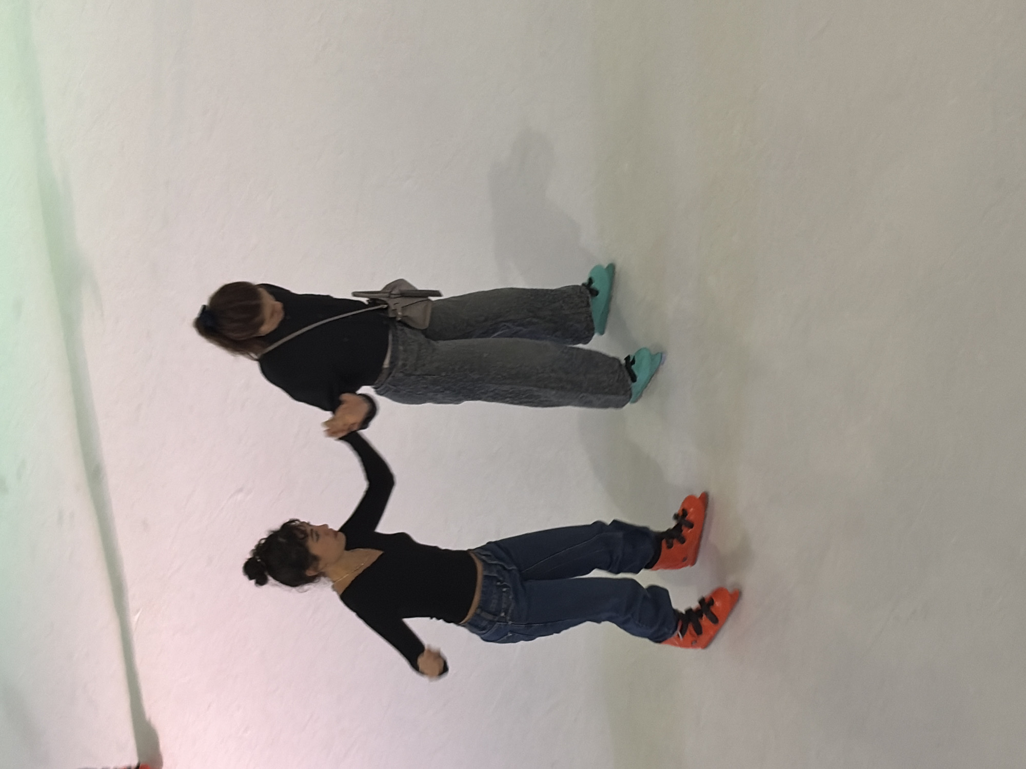 This image captures two young women ice skating at an indoor ice rink in Thủ Đức, Vietnam. The woman on the left, possessing dark, curly hair styled in a high bun, wears a black long-sleeved V-neck top and dark blue baggy jeans, completing her attire with bright orange ice skates featuring black laces. She looks towards the right, extending her left arm to hold hands with the second woman. The woman on the right has dark hair secured in a ponytail with a dark scrunchie and is looking down at the ice. She wears a black long-sleeved top, grey loose-fitting pants, and light teal ice skates with black laces, carrying a small grey crossbody bag over her left shoulder. Both individuals are actively engaged in skating, with the hand-holding indicating an interaction of support or shared movement. The environment consists of a smooth, white ice surface with visible skate marks, illuminated by overhead lighting that produces a faint greenish tint in the upper portion of the frame and subtle shadows beneath the skaters. A portion of a third person's red skate is visible in the upper left corner.