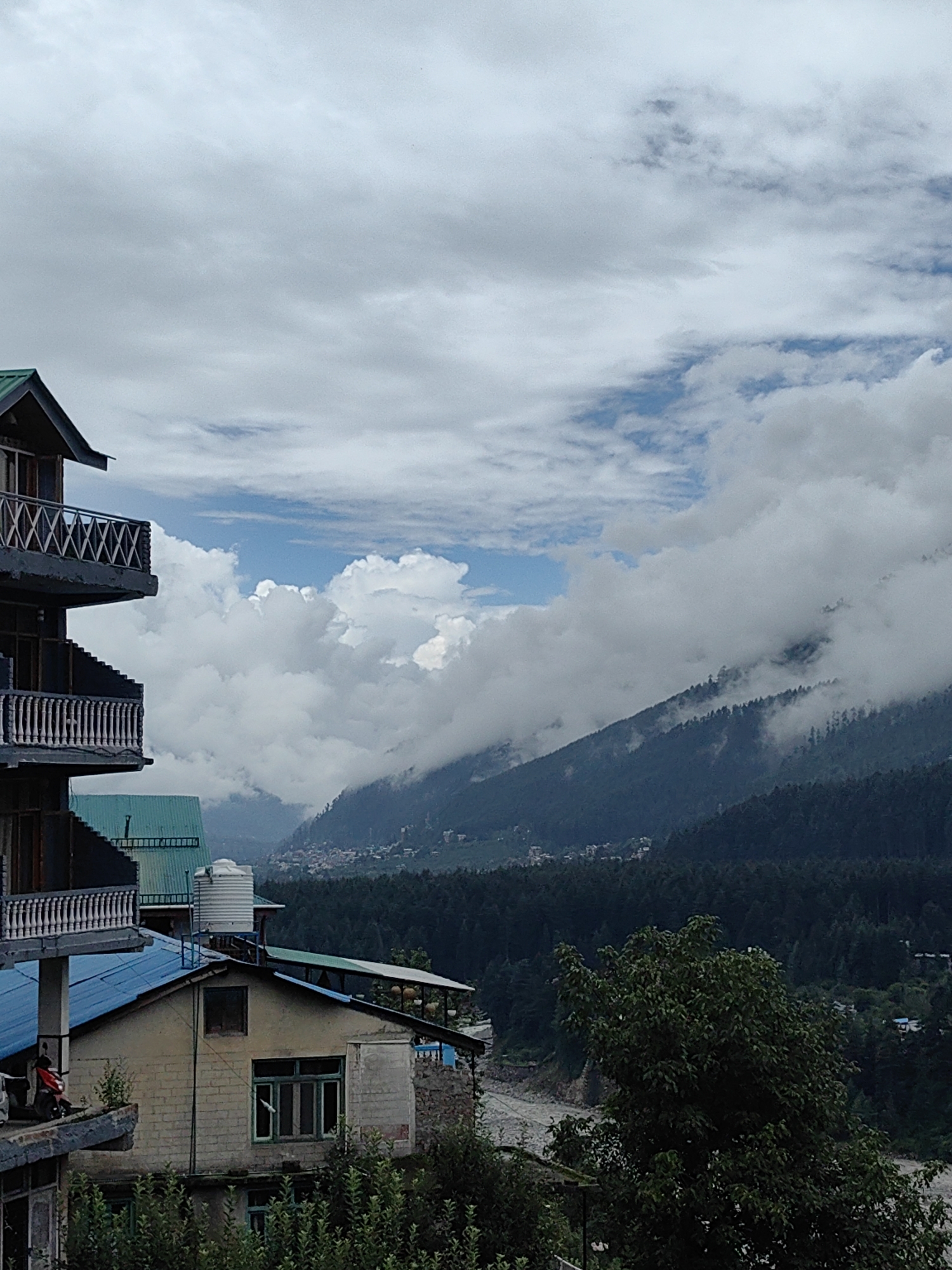 The image depicts a scenic mountain landscape with several buildings in the foreground, likely in or near Vashisht, India. The sky is partly cloudy with dramatic formations of white clouds interspersed with patches of blue.
In the left foreground, a multi-story building with a dark exterior and white balustrades on its balconies is visible. Below this, another building with a blue roof is partially seen, and further down, a lighter-colored building with a green-framed window is prominent. This building has a water tank on its roof and appears to be constructed of brick. A scooter is parked on a ledge in front of one of the lower buildings. Lush green foliage and trees surround the buildings and extend into the middle ground.
The background is dominated by rolling hills covered in dense, dark green pine forests. These hills recede into the distance, with some appearing to be partially shrouded in clouds. A faint glimpse of a settlement or village can be seen nestled in the valley between the forested hills. A river or water body is barely visible at the bottom center of the image, flowing through the valley. The overall atmosphere is one of natural beauty and a rural or semi-rural setting.