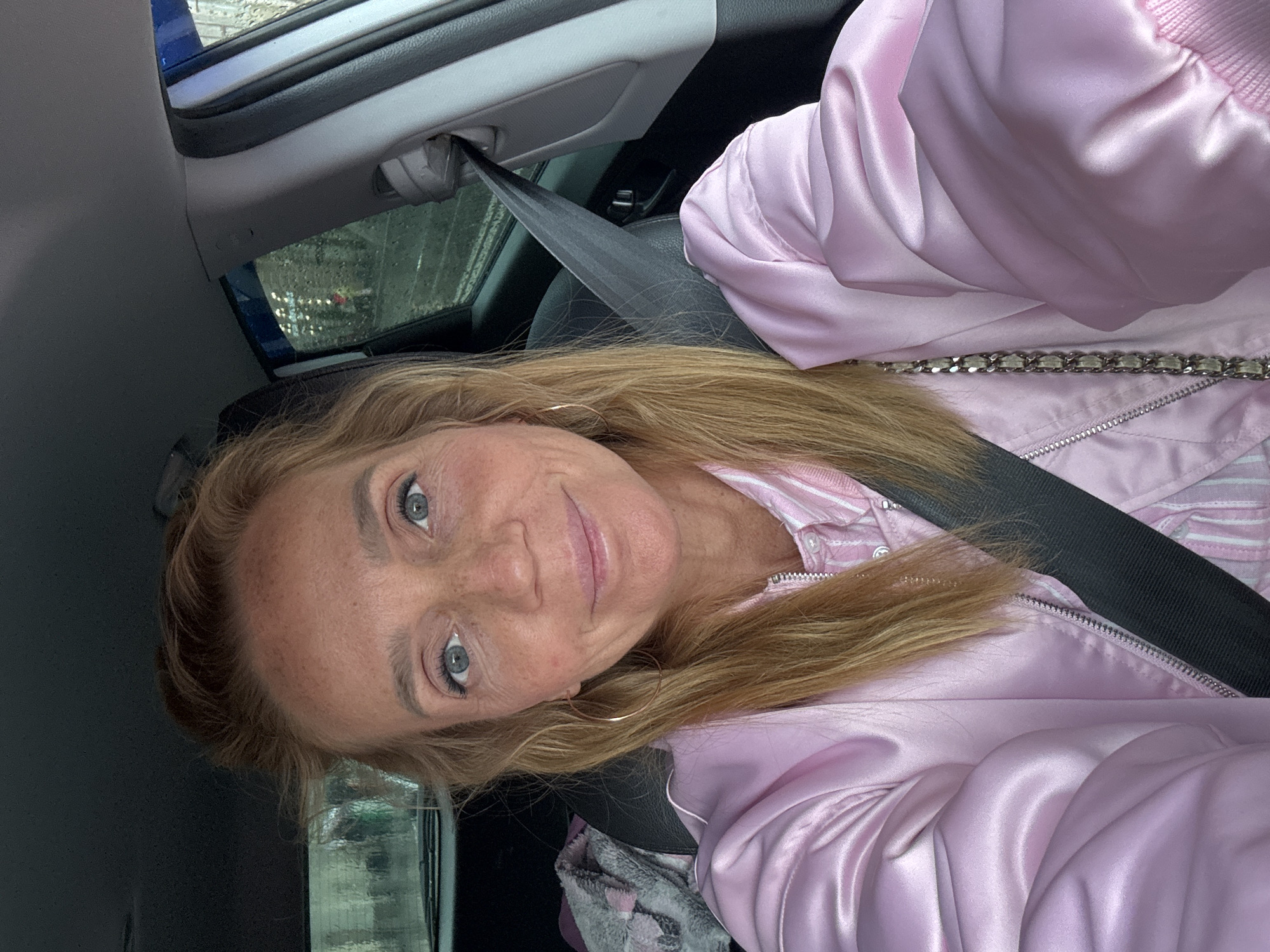 A woman with blonde hair and blue eyes, wearing a pink satin bomber jacket over a pink and white striped shirt, is taking a selfie in a car. Her seatbelt is fastened. In the background, through the car window, are reflections of lights and possibly buildings. A gray blanket with a pink pattern is visible on the passenger seat. The lighting suggests an overcast day. There are no identifiable landmarks that place this image specifically in Saint Petersburg, Russia.