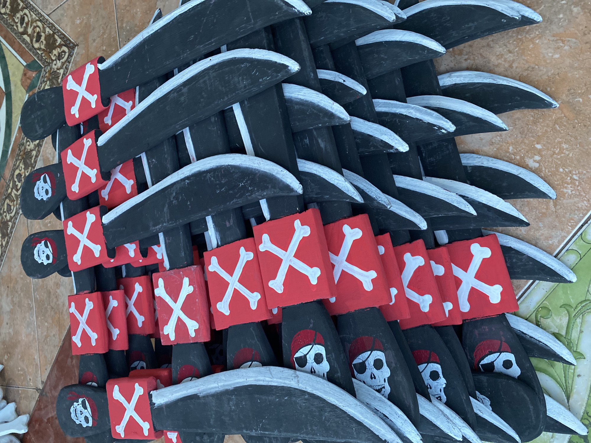 A high-angle, close-up shot depicts a stack of numerous toy pirate swords on a patterned tile surface. The swords have black blades edged with white paint, handles adorned with red squares bearing white skull-and-crossbones designs, and skull-and-crossbones painted at the base of the handles. The scene is located in Tegallalang, Indonesia.