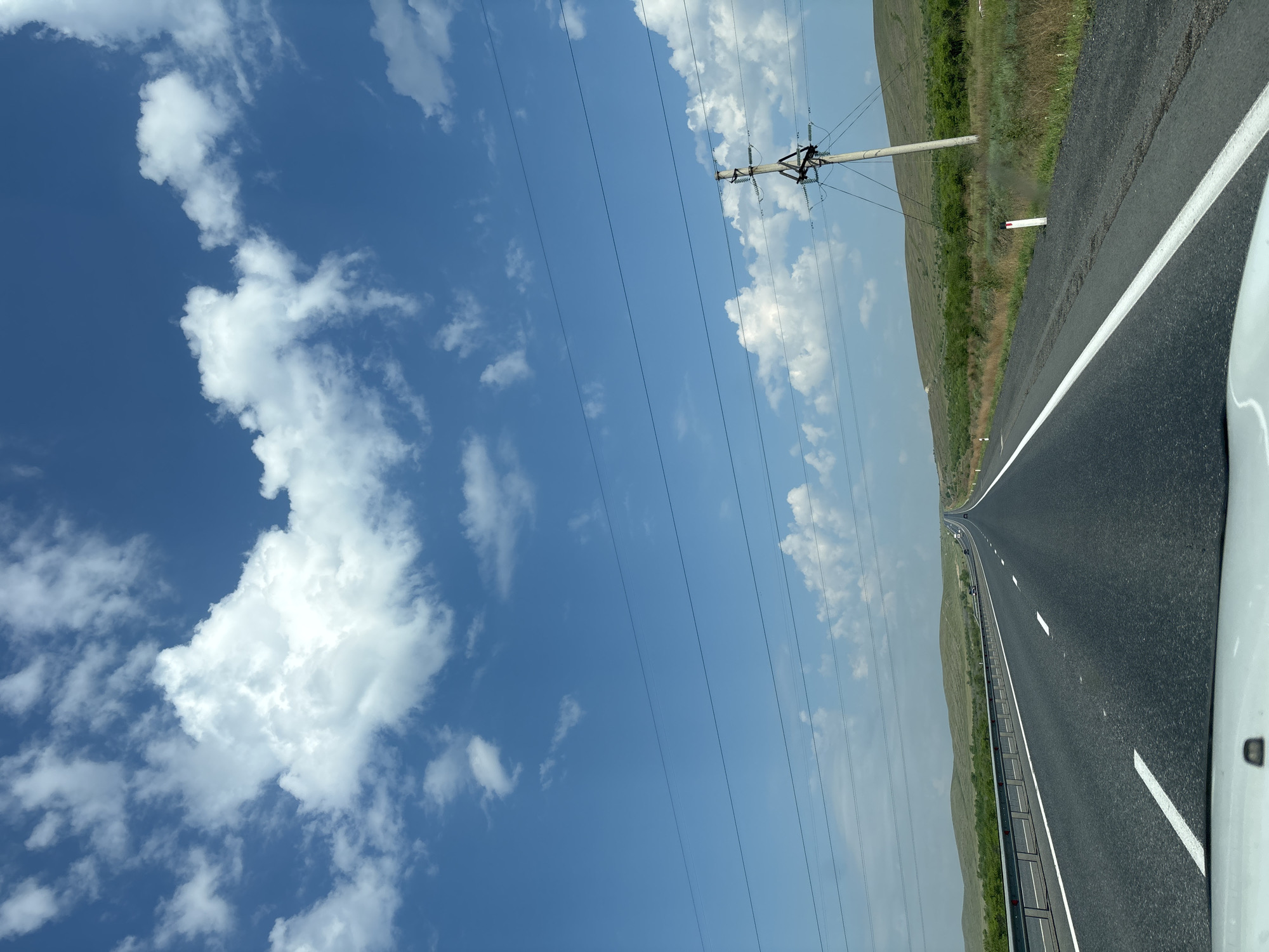 The image depicts a view from a vehicle driving on a paved highway under a bright blue sky with scattered white clouds. The road stretches into the distance, bordered by a metal guardrail on the left and a grassy, undulating landscape on the right. Power lines are visible overhead, supported by concrete poles. The terrain appears rural, with low hills covered in green vegetation and scattered trees. There are no people visible in the image, and no specific landmarks are identifiable that would indicate the location in Chernogorsk, Russia. The overall scene is one of travel on an open road in a natural environment.