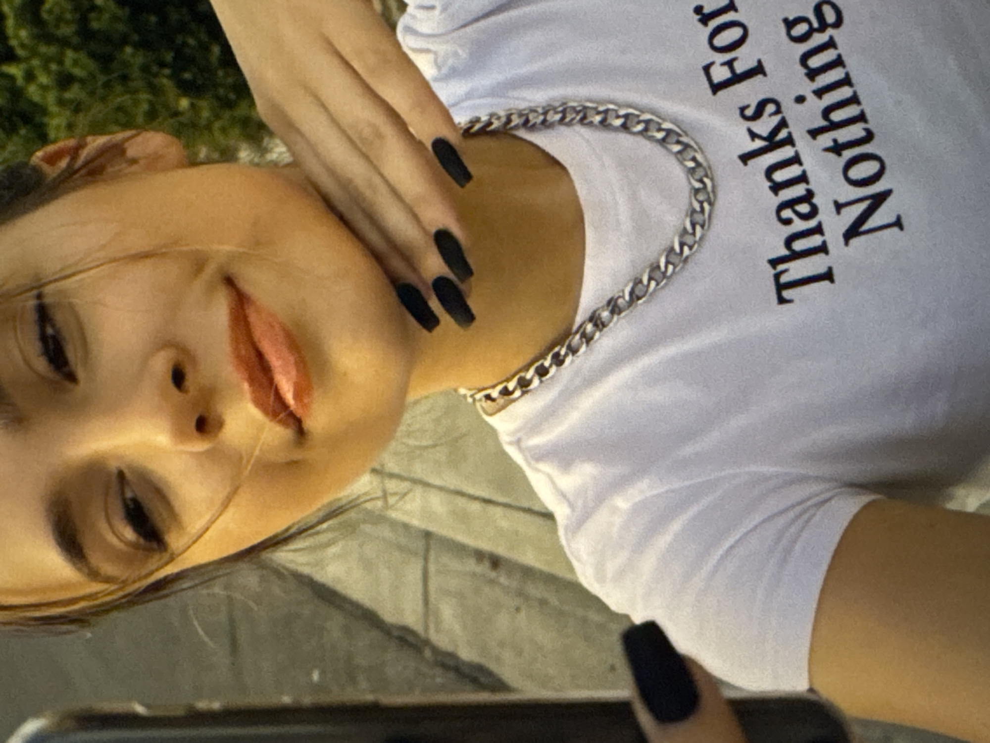 The image shows a close-up of a young woman taking a selfie outdoors. She is wearing a white t-shirt with black text that reads "Thanks For Nothing". She has black painted nails and is wearing a silver chain necklace. Her hand is resting on her neck, and she is smiling slightly. The background is blurred, but appears to be a street scene with some greenery. The specific location in Kaunas, Lithuania is not discernible from the image.