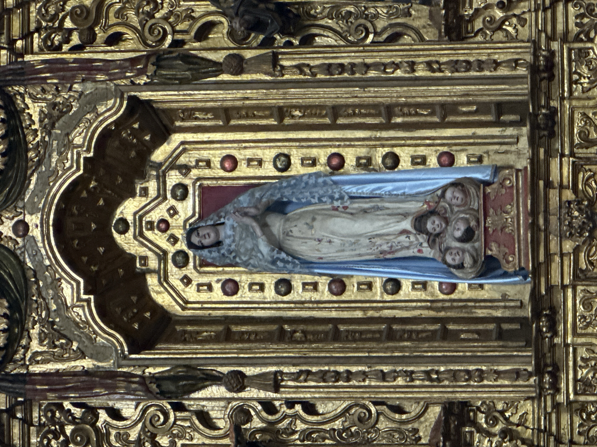 This image features a highly ornate, gilded religious sculpture, likely a section of a Baroque altarpiece or retablo, characteristic of churches in Mexico City, Mexico. At its center, a polychromed statue of a recumbent female figure lies peacefully within a deep, maroon-red niche. The figure, possibly depicting the Virgin Mary or a saint, is dressed in a light-colored gown adorned with subtle floral patterns and draped in a pale blue cloak. A delicate, light-colored veil covers her head, and her expression is serene and contemplative. Clustered around her feet are several smaller, idealized heads, resembling cherubs or putti.
The statue is surrounded by an exceptionally elaborate gilded wood frame, showcasing intricate carvings, swirling volutes, geometric motifs, and raised architectural embellishments. Dark red and brownish-gold spherical ornaments are embedded at regular intervals along the gilded borders of the niche, enhancing the rich detailing. The meticulous craftsmanship of the gold leaf and carved wood creates an impression of grandeur and opulence. The scene evokes a sense of reverence and devotion, typical of colonial-era religious art. No text is visible in the image.