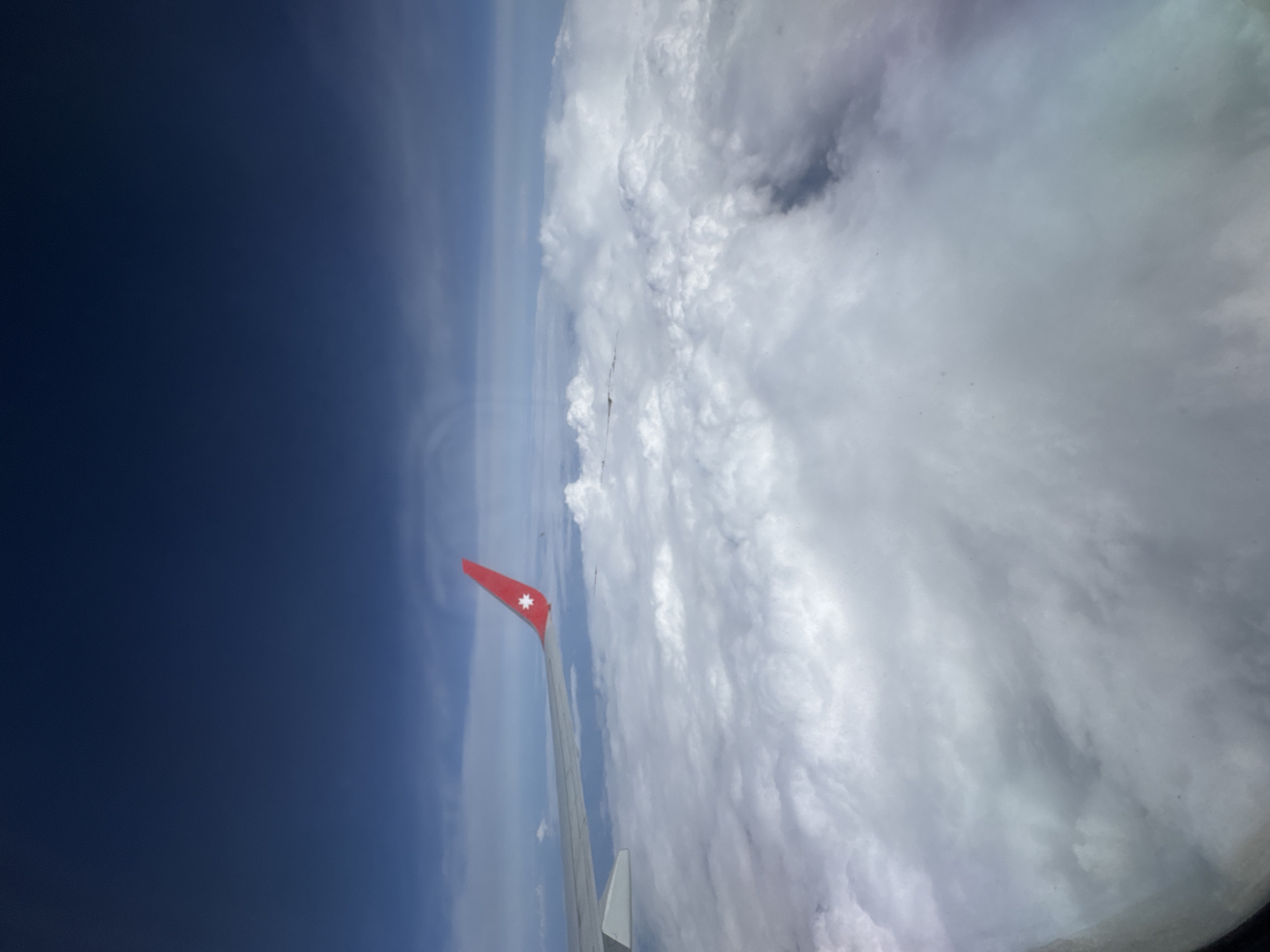 The image shows a view from an airplane window. The wing of the plane is visible in the lower left, painted with a red tip and a white star-like emblem. To the left of the wing is a clear blue sky that fades into a lighter blue as it moves away from the wing. To the right of the wing is a dense mass of swirling white clouds. The image provides no information to confirm the location of Lyamtsino, Russia. There is no information to confirm information about people or events.