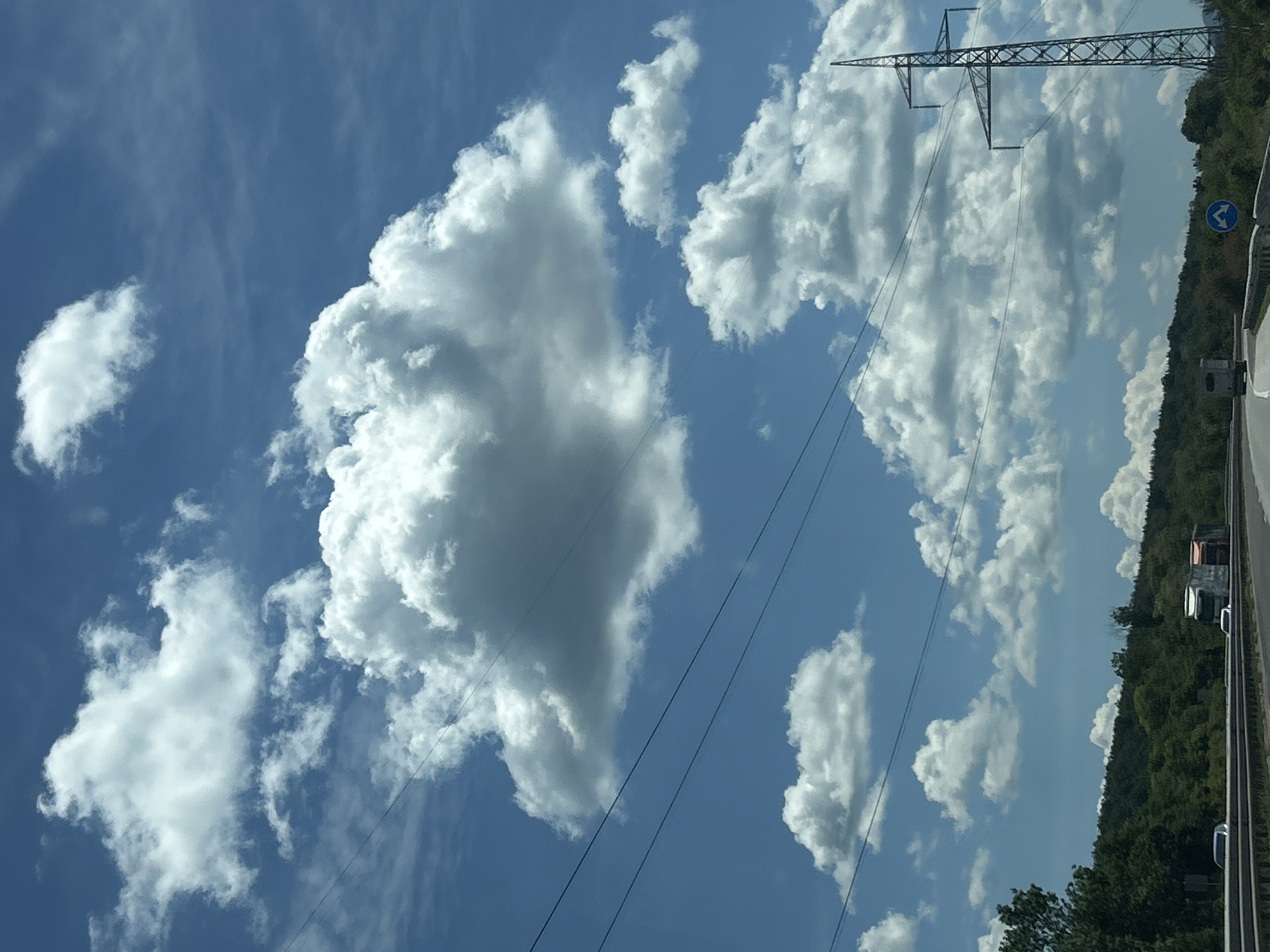 The image depicts a cloudy sky above a highway in the vicinity of Verinsko, Bulgaria. The sky is a vibrant blue, dotted with numerous white, fluffy cumulus clouds of varying sizes. Several power lines stretch horizontally across the frame, obscuring parts of the sky and landscape. On the right side of the image, a tall, metallic electricity pylon stands prominently against the clouds.
Below the sky, a tree-covered hillside forms the horizon. The highway is visible in the foreground, with a few vehicles including cars and trucks. A traffic sign with blue arrows indicating a diversion is also visible on the right side of the highway. The overall scene suggests a clear day with scattered clouds, likely captured from a moving vehicle on the highway.
