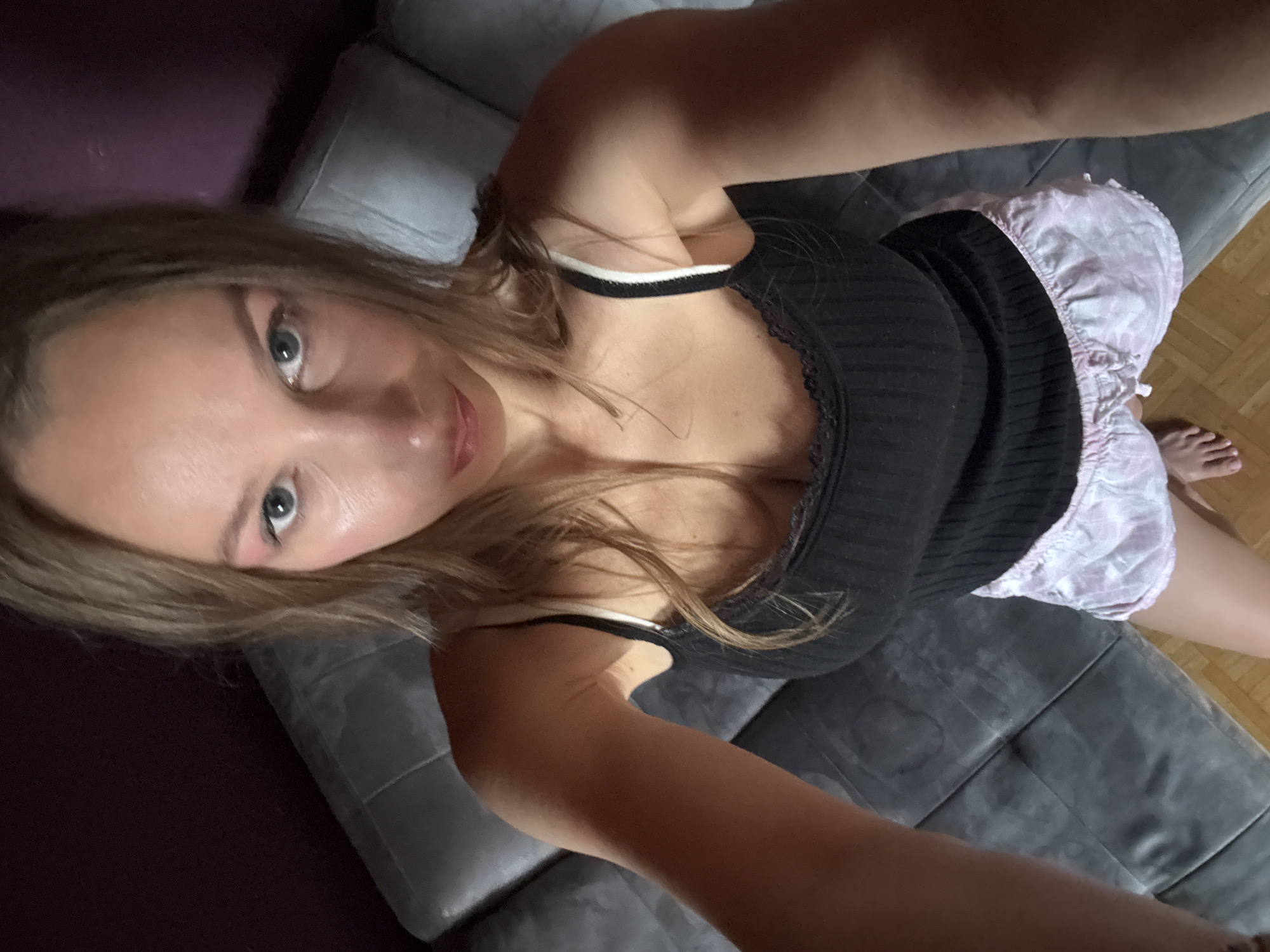 A woman is sitting on a gray sofa with a dark purple wall behind her and a wooden floor in front of her. She is wearing a black tank top with white trim and pink patterned shorts. She has long blonde hair and is wearing makeup. The photo is taken from a high angle, looking down at the woman. The location is not visible in the image.