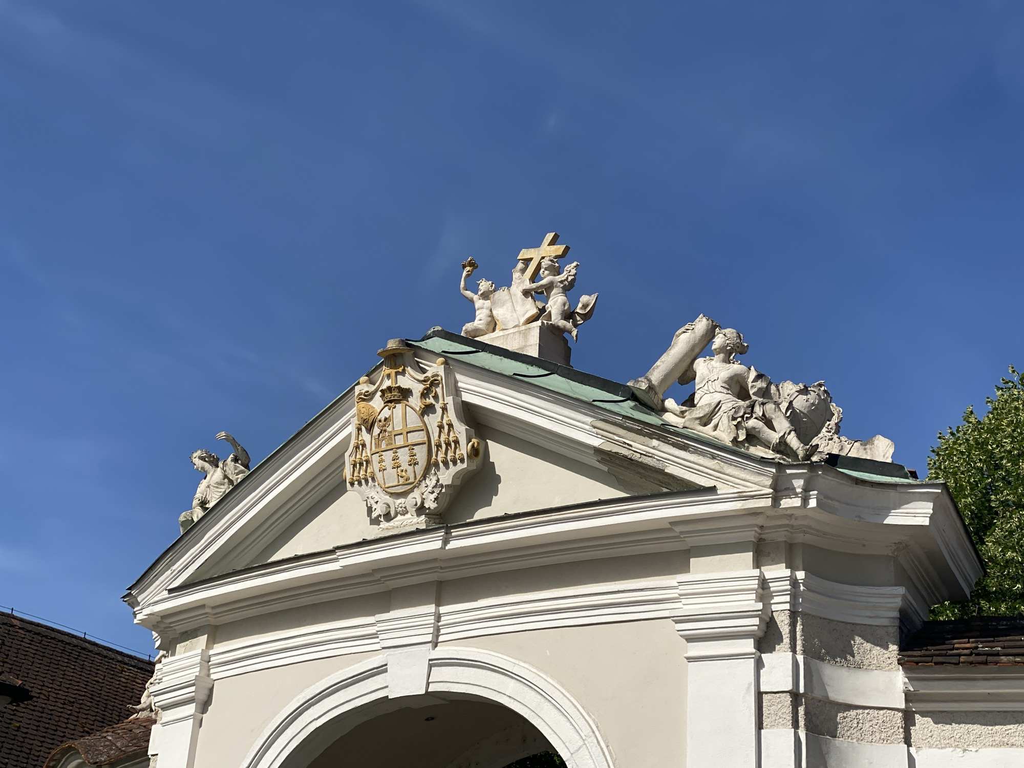 The image shows the top of an ornate gateway in Sankt Pölten, Austria. The gateway is topped with several classical sculptures against a clear blue sky. Prominently featured is a coat of arms in gold and yellow, located on the pediment of the gateway, which is topped with a green roof. Above the pediment, a cross is being held up by two cherubic figures. To the right, a larger sculpture depicts a female figure holding a large cylindrical object. To the left of the gateway's pediment, another sculpture of a seated figure is visible. The architectural style suggests a historical or ecclesiastical building.