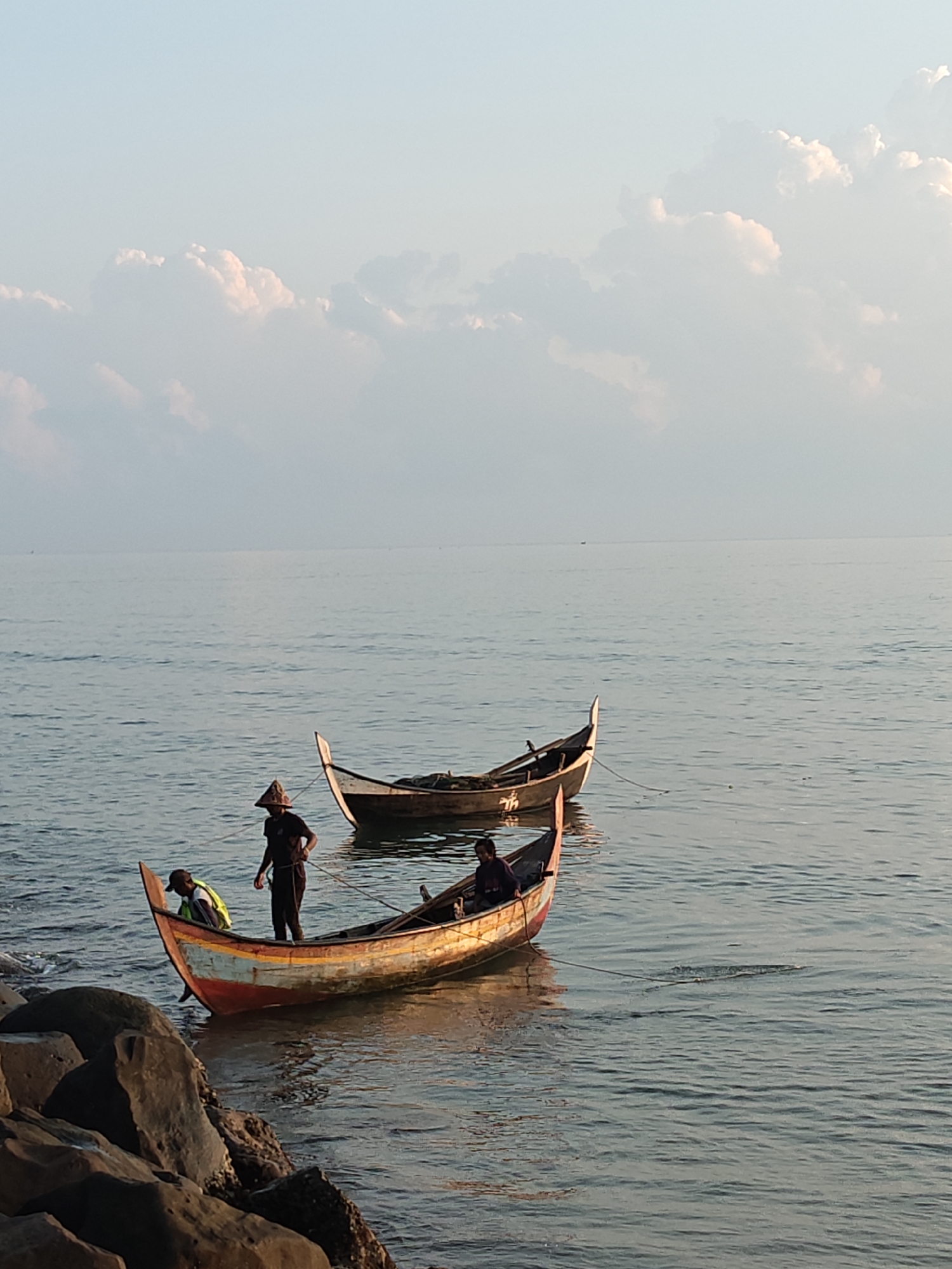 Two outrigger boats float on the calm sea near a rocky shore in Lhokseumawe, Indonesia. Three people are on or near the boat closest to the shore. One person wears a yellow vest and baseball cap, and is bent over in the front of the boat. A second person stands holding a rope, wearing a hat, and dark clothing. A third person sits at the rear of the boat. The second boat is further out in the water, and no persons are visible in it. The sky is clear, with fluffy clouds.