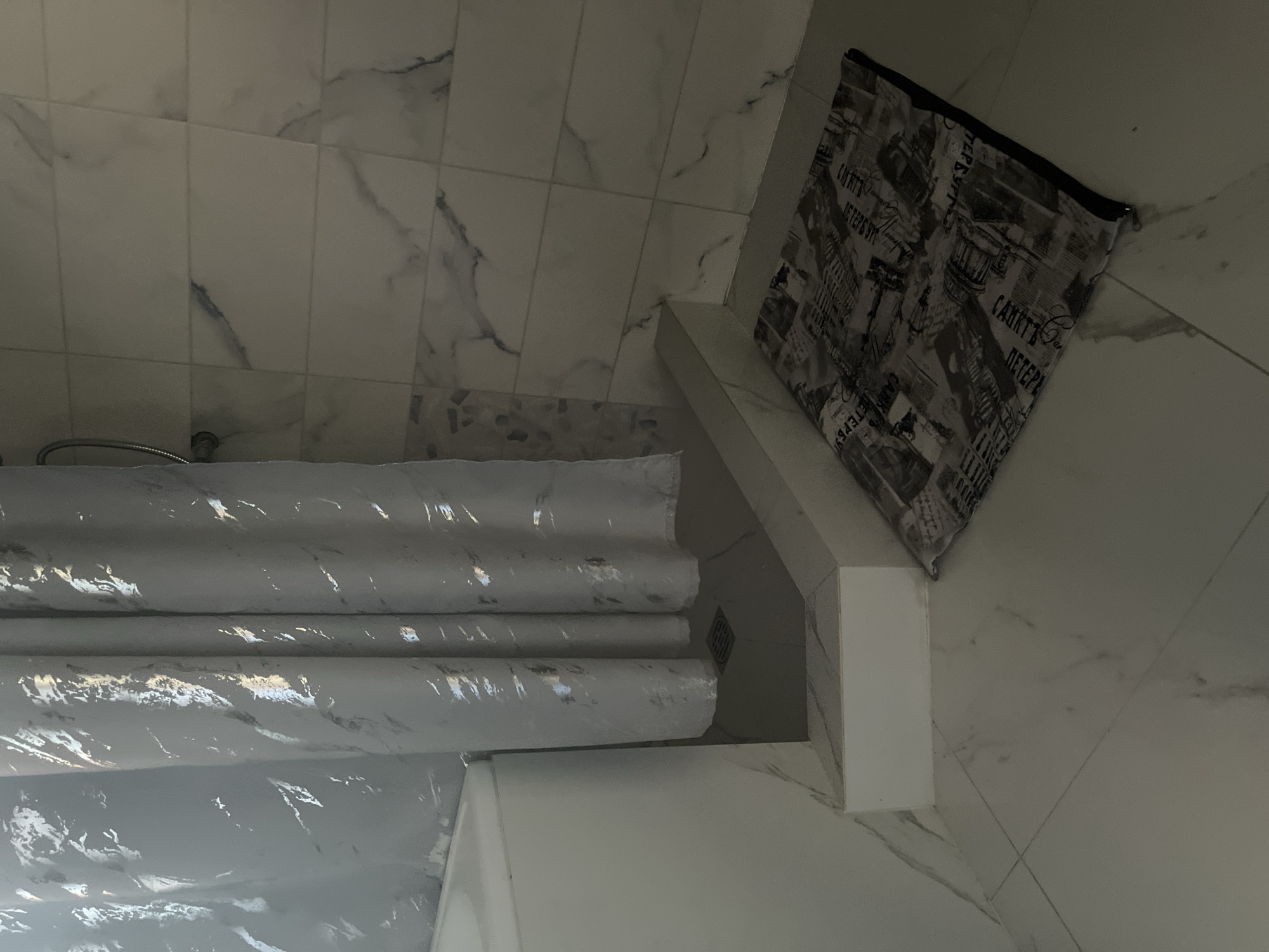 The image depicts a corner of a bathroom, likely in Saratov, Russia. A white bathtub is visible on the left, partially obscured by a grey and silver patterned shower curtain. The shower curtain hangs over a tiled shower stall, which has a dark drain visible on the floor. The walls of the shower are tiled with large, light-colored tiles featuring subtle grey veining. A patterned bath mat, with a design resembling newsprint or old text, lies on the tiled floor outside the shower. The overall impression is of a modern, clean bathroom interior. There are no people or discernible actions taking place in the image.