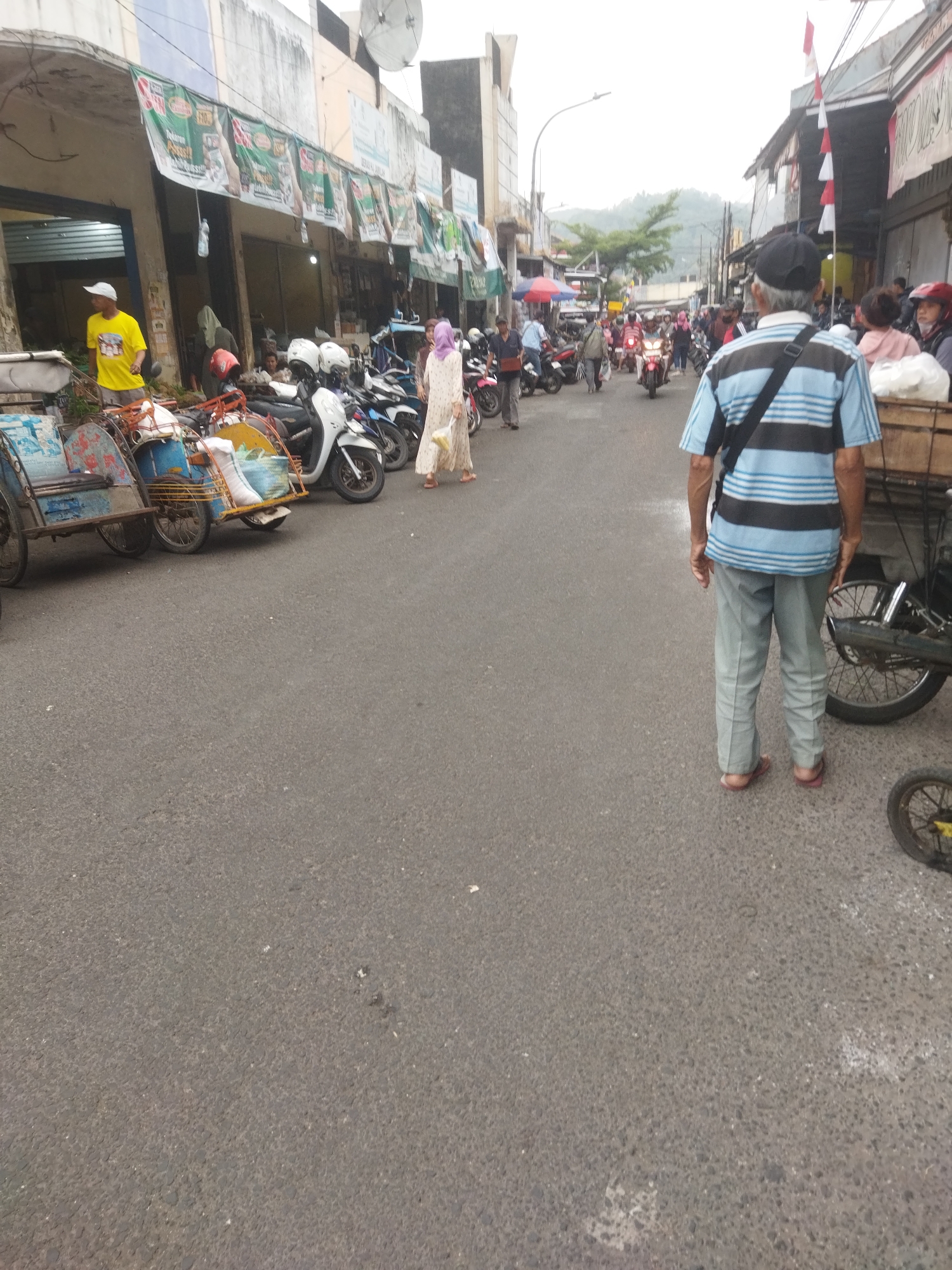 A street scene in Banjar, Indonesia, shows a row of parked motorcycles and several pedicabs on the left side of the road, suggesting a commercial or market area. In the center of the image, people are walking along the street, some wearing traditional Indonesian clothing. A man in a blue and black striped shirt and light gray pants is walking away from the camera on the right side of the road, carrying a black bag over his shoulder. Behind him, a cart loaded with goods is partially visible. The street appears to be paved with asphalt, and buildings line the left side of the road, displaying banners and advertisements. The sky is overcast, and a hilly landscape is visible in the background. The overall impression is of a busy, everyday street scene in an Indonesian town.