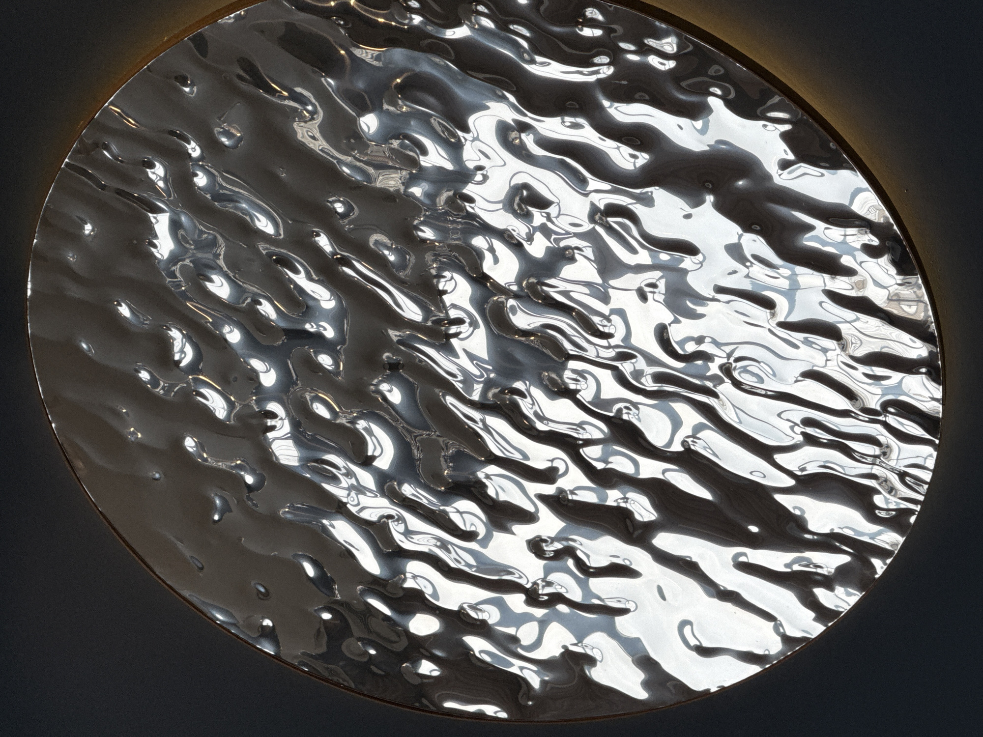 A close-up shot shows a circular, textured, silver-colored metal plate. The surface of the plate is uneven, creating a pattern of light and shadow. The plate is mounted against a dark gray background. A warm light emanates from behind the plate, illuminating its edge. There is no indication of a location in Votkinsk, Russia, or any people, actions, or interactions.