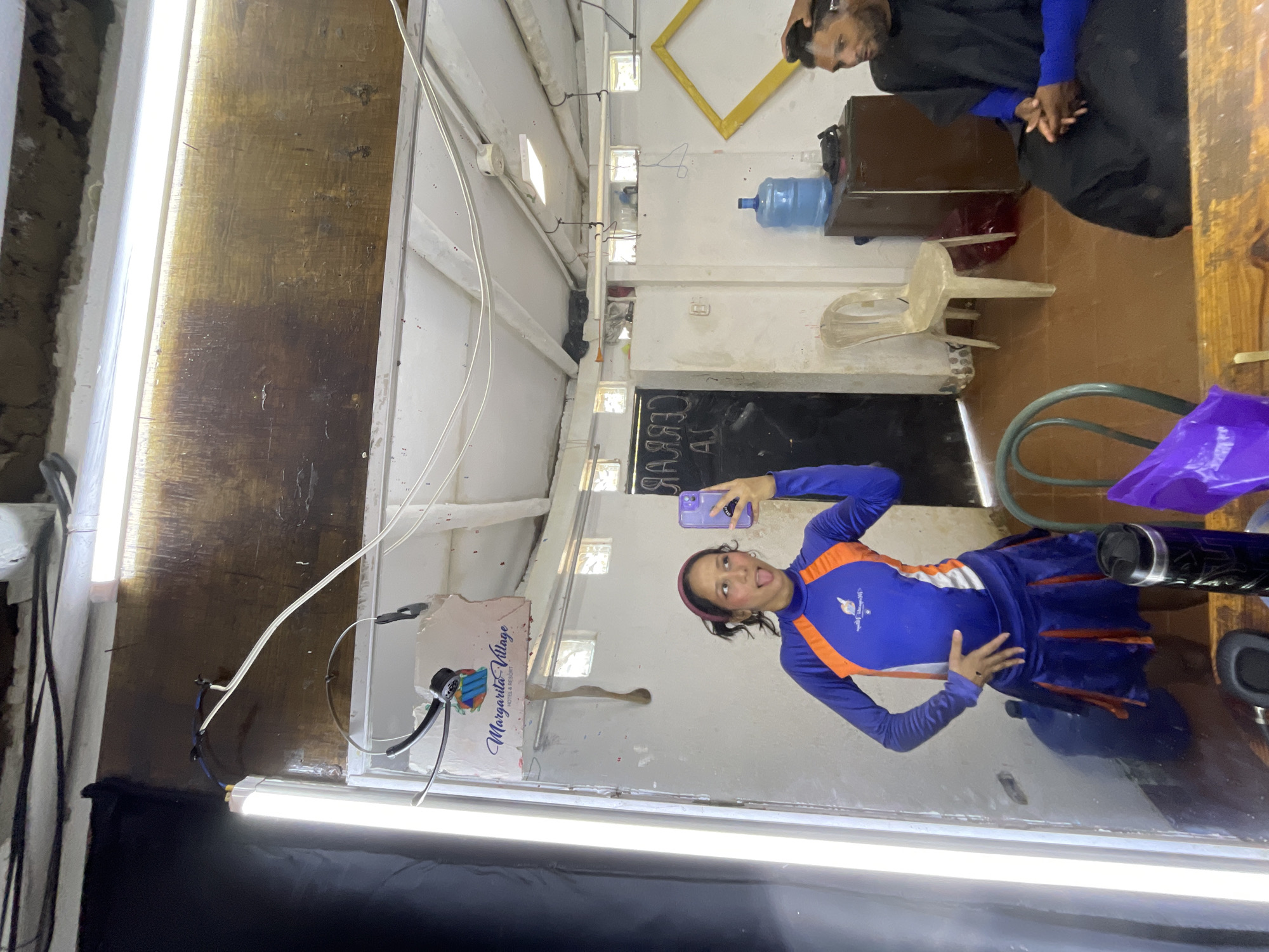 A young woman with dark hair pulled back and wearing a purple headband, sticks her tongue out at the camera as she takes a selfie in a mirror. She is wearing a blue and orange long-sleeved athletic top and a matching pleated skirt. The background appears to be a dimly lit, cluttered room. The room has a wooden ceiling with exposed wires and fluorescent lighting. A sign for "Margarita Village Hotel & Resort" hangs on the wall. To the right, a man with dark skin is seated and appears to be getting his hair cut, covered with a black cape. There is a large blue water dispenser, a brown cabinet, and several chairs visible in the room. The overall setting suggests a backstage area or a utility room within a resort in Porlamar, Venezuela.