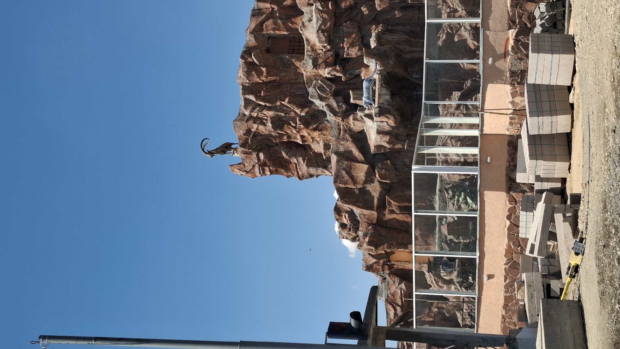 The image captures a zoo exhibit designed to mimic a rocky mountain environment under a clear blue sky in Shymkent, Kazakhstan. In the foreground, stacks of concrete blocks and paving stones indicate ongoing construction or landscaping work. A traffic light pole stands to the left. The main focus of the exhibit is a large, man-made rock formation, resembling a cliff face. Perched precariously on the highest point of this rock formation is a statue of an ibex, a wild goat known for its long, curved horns. Below the ibex statue, on a lower ledge of the rock formation, is a statue of a snow leopard. The rock formation features carved details and a small doorway. A glass-enclosed viewing area is visible behind the rock formation, allowing visitors to observe the exhibit. The zoo itself appears to be undergoing some renovations or development, suggested by the construction materials in the foreground.