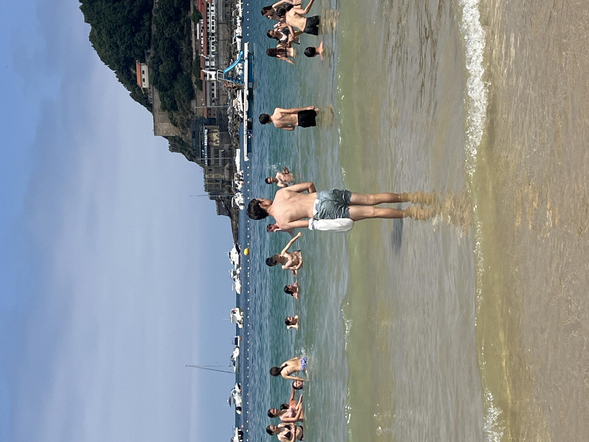 A group of people, primarily young individuals, are enjoying a sunny day at La Concha Beach in San Sebastián, Spain. The scene captures the vibrant atmosphere of the popular tourist destination. In the foreground, a young boy stands facing away from the camera, partially submerged in the shallow, clear, turquoise water. His back is to the viewer, and he wears light-colored shorts and has a white bag slung over his shoulder. Behind him, a diverse group of people are scattered throughout the water. Some are interacting with each other, while others are simply swimming or wading. The water is calm and inviting, with small waves gently lapping at the shore.
In the mid-ground, several boats are anchored in the bay, their white hulls and masts creating a picturesque backdrop. Beyond the boats, the distinctive coastline of San Sebastián is visible. The lush green slopes of Monte Urgull rise dramatically from the sea, topped with a historic fortress. The buildings of the city are nestled along the hillside and at the base of the mountain, painted in various shades of white, cream, and terracotta, adding to the charming aesthetic of the location. The sky is a clear, bright blue, indicating a perfect summer day for beach activities. The overall impression is one of leisure and enjoyment in a beautiful coastal city.