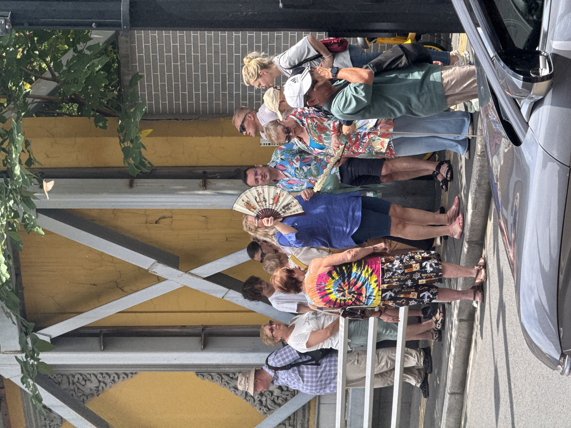 The image captures a daytime scene in what appears to be an urban environment, likely in the Jing'an District, China. A group of approximately a dozen people, ranging in age, are standing in a line or queue on a paved walkway.
The individuals are dressed in casual summer clothing, including shorts, t-shirts, light blouses, and sandals, suggesting warm or hot weather. Several people are holding or actively using fans to cool themselves; one person in a blue top is seen fanning themselves with a decorative, traditional Chinese-style fan adorned with painted figures. Other notable attire includes a brightly colored tie-dye top and patterned black shorts with white anchor motifs and the word "SAILOR" repeated. Many wear sunglasses.
To the right, the front fender and headlight area of a silver or grey car are visible, parked alongside the curb. The background features a building with a textured yellow wall, showing signs of wear and cracking, and above it, a section of a grey brick wall with white mortar. Green foliage from trees or vines cascades down from above the structures on the left side of the image. Metal beams and decorative architectural elements are also present, forming part of an overhead structure or building facade. The bright lighting and distinct shadows indicate it is daytime. The people appear patient, and the overall mood is calm.