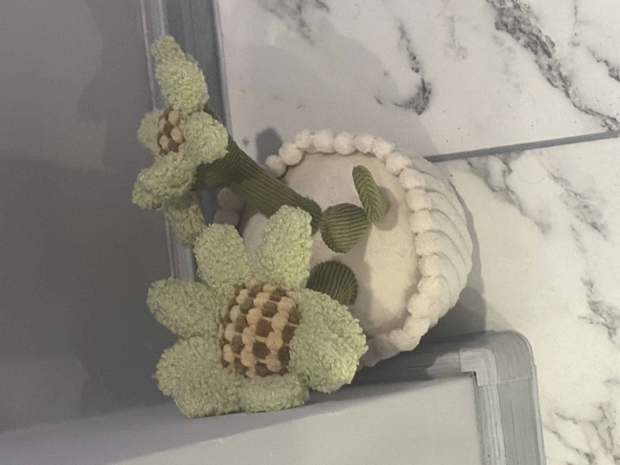 A fuzzy, light green, potted artificial flower with a brown and tan checkered center sits on a marble-patterned floor. The pot is white and textured with a ruffled edge and appears to be made of a plush material. The flower is placed in a corner, with a light gray wall on the left and the floor extending to the right. The scene is indoors, with soft lighting. No people or actions are visible. The image is an indoor scene, likely a domestic setting, with no discernible connection to the city of Podgumer, Bulgaria.