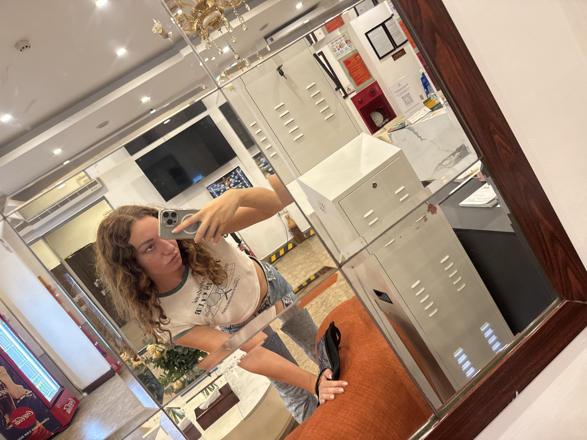 The image is a mirror selfie taken by a young woman with curly brown hair. She is wearing a white cropped t-shirt with a tennis graphic and light-wash denim shorts. She is holding a smartphone in her right hand, angled to capture her reflection. In the reflection, the setting appears to be indoors, possibly a hotel lobby or a common area. There is a large mirrored wall, reflecting various objects and parts of the room.
Visible elements in the reflection include:
* A dark-framed mirror, with a wooden finish on the right side.
* A large screen television mounted on the wall.
* A Coca-Cola vending machine to the left.
* What appear to be lockers or storage cabinets.
* A red fire extinguisher mounted on the wall, with safety signs above it.
* A patterned floor with yellow and black hazard stripes.
* A plush orange couch or seating area where the woman's legs are resting.
* A black face mask is placed on the couch.
* A ornate chandelier with crystals hangs from the ceiling.
* Fluorescent lighting is visible in the ceiling.
The overall impression is casual and relaxed. The lighting suggests it could be daytime, as there is ample illumination. The presence of the vending machine and the general decor hint at a public space, possibly a hotel or a service area. There is some visible text on the vending machine, which appears to be in a non-Latin script. Also, a QR code is visible on a white sign in the reflection. The woman's expression is playful, as she puckers her lips for the selfie.