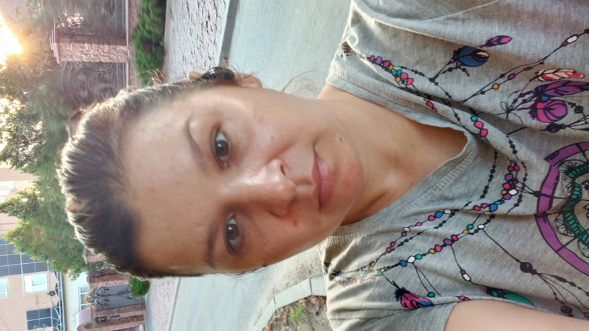 A close-up, selfie-style photograph captures a woman with dark hair pulled back. She is wearing a gray t-shirt with a dreamcatcher design. The background suggests an outdoor urban setting, with a building and trees visible. The sun is setting, casting a warm glow. The precise location within Kropyvnytskyi, Ukraine cannot be determined from the image alone.