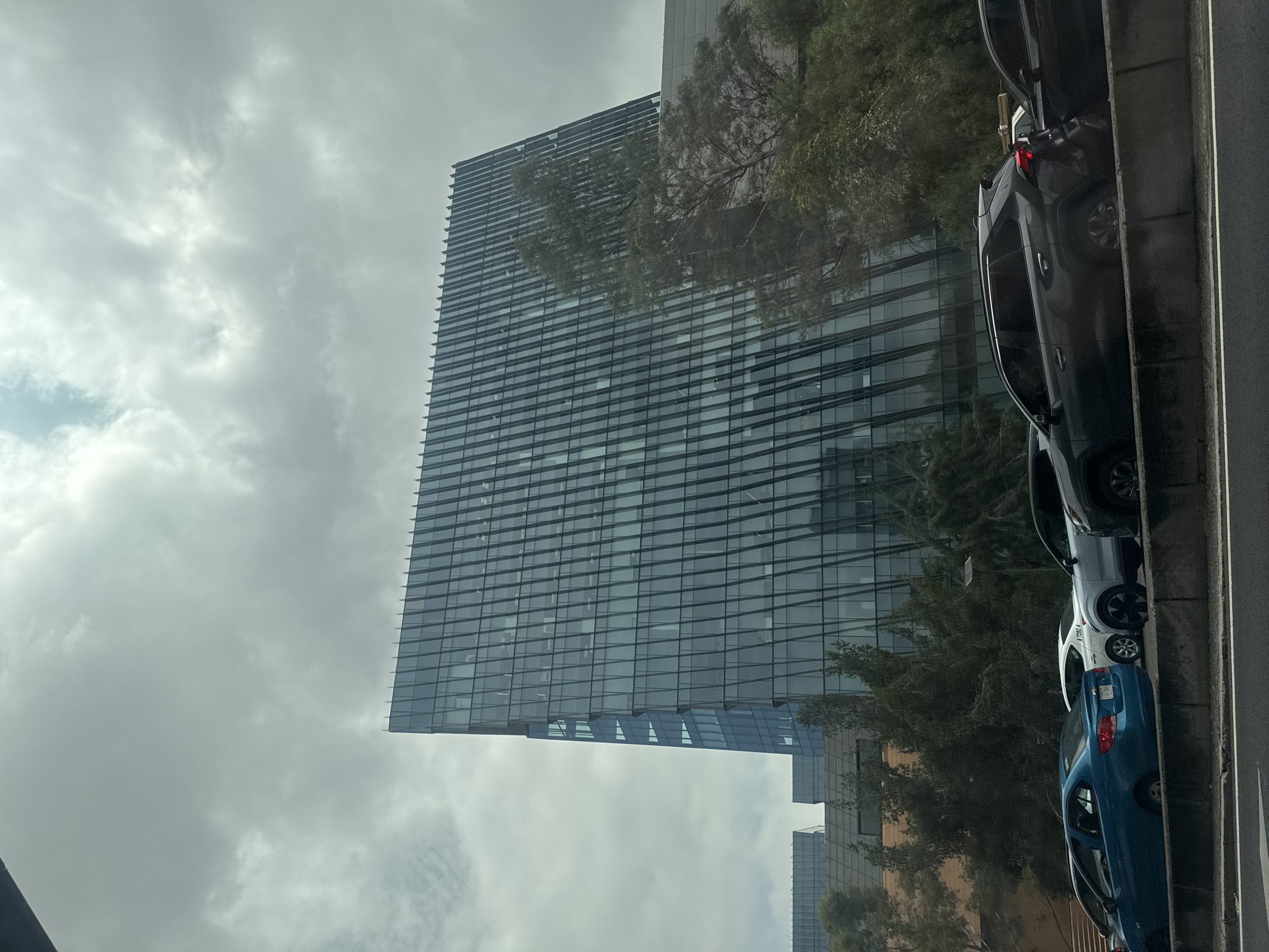 The image shows a tall, modern skyscraper with a glass facade, reflecting the overcast sky. The building is located in Mexico City, Mexico. The image is taken from street level, with several parked cars visible along the right side of the frame. The cars are various colors: black, silver, and blue. Trees partially obscure the base of the building, adding some greenery to the urban scene. The sky is filled with grey clouds, contributing to a muted, slightly somber atmosphere. The perspective of the image is from a lower angle, emphasizing the height of the skyscraper.