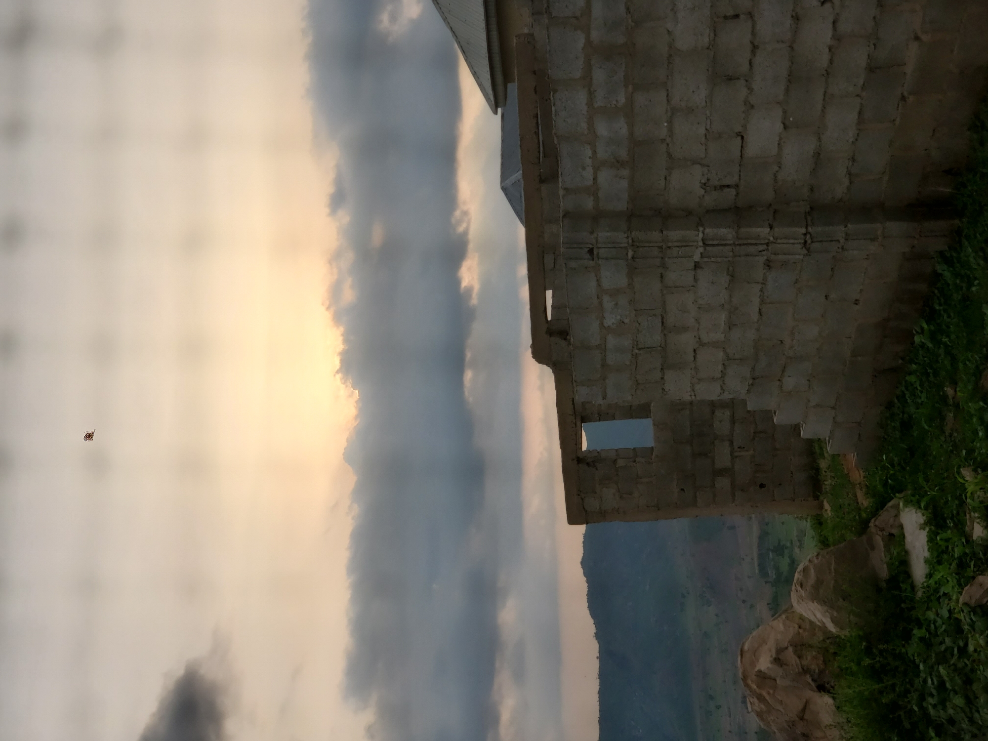 A partially constructed cinder block building stands in the foreground, its walls incomplete and openings visible. To the left, large rocks and green vegetation sit on a rocky outcrop. In the middle ground, a landscape of rolling hills and greenery stretches towards a distant mountain range. The sky above is filled with dramatic, overcast clouds, with rays of sunlight breaking through near the horizon, suggesting either sunrise or sunset. A small, dark object, possibly a kite or bird, is visible in the upper portion of the sky. The scene appears to be set in a rural or undeveloped area.