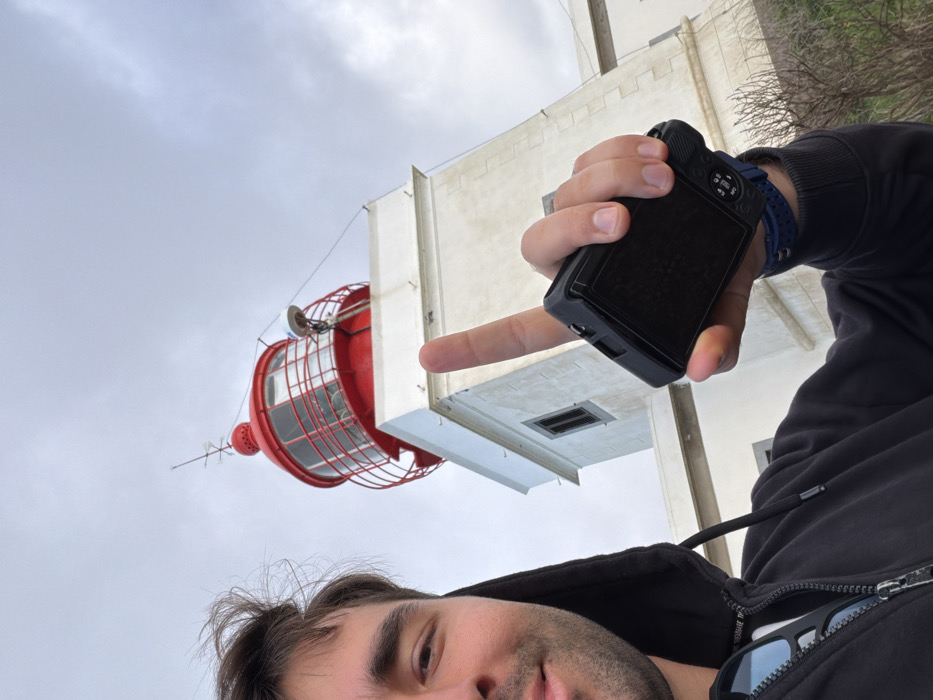 The image captures a unique selfie taken from a low, tilted perspective, featuring a man and a distinctive architectural structure against a cloudy sky.
In the bottom right portion of the image, the upper part of a man's face is visible, including his right eye, eyebrow, forehead, part of his nose, and mouth, along with stubble on his chin and cheek. He has dark hair, styled to be somewhat messy. He is wearing a dark, possibly black or navy, hooded top with a zipper down the front. A pair of sunglasses is hooked onto the front of his clothing, near the zipper.
The man's right hand dominates the central-right area of the image. He is holding a small, black digital camera, possibly a mirrorless or advanced compact model, with its back facing the viewer. The camera has a screen and visible buttons/dials. His index finger is extended, pointing directly towards a light-colored building in the background. A blue, textured wristband is visible on his right wrist.
In the background, to the left and center, a prominent red structure resembling the upper part of a lighthouse is visible. It features a cylindrical or slightly conical red base topped with a large, red, cage-like structure enclosing a glass-paneled lantern room. Below the main red section, there are several antennae or communication devices extending downwards. Behind and to the right of this red structure, a tall, light-colored (likely white or off-white) building or tower rises. This building exhibits modern, angular architecture with multiple horizontal and vertical structural elements, including what appear to be vents or square openings on its surface.
The sky overhead is overcast and light grey, indicating a cloudy or muted day. The overall perspective is looking upwards, suggesting the man is positioned below the structures he is photographing. The man's gesture of pointing his finger towards the building creates a playful or intentional visual connection between himself and the distant landmark.