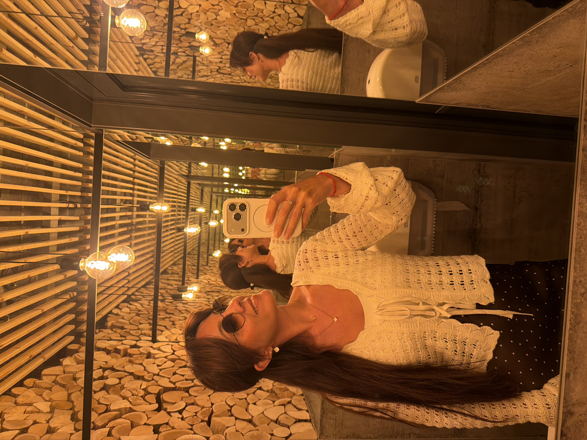 A woman is taking a mirror selfie in what appears to be a restroom. The walls of the restroom are decorated with a mosaic of cut wood logs, and the ceiling features exposed wooden beams and light bulbs. The woman has long dark hair, wears round sunglasses, a white knit cardigan over a white top, and a dark polka-dot skirt. She is wearing jewelry, including earrings and necklaces, and a red bracelet on her right wrist. In her right hand, she holds a smartphone, capturing her reflection. The mirror reflects multiple instances of the woman and the restroom's interior. The overall lighting is warm and ambient, contributing to a rustic yet modern aesthetic. The location is identified as Riga, Latvia.