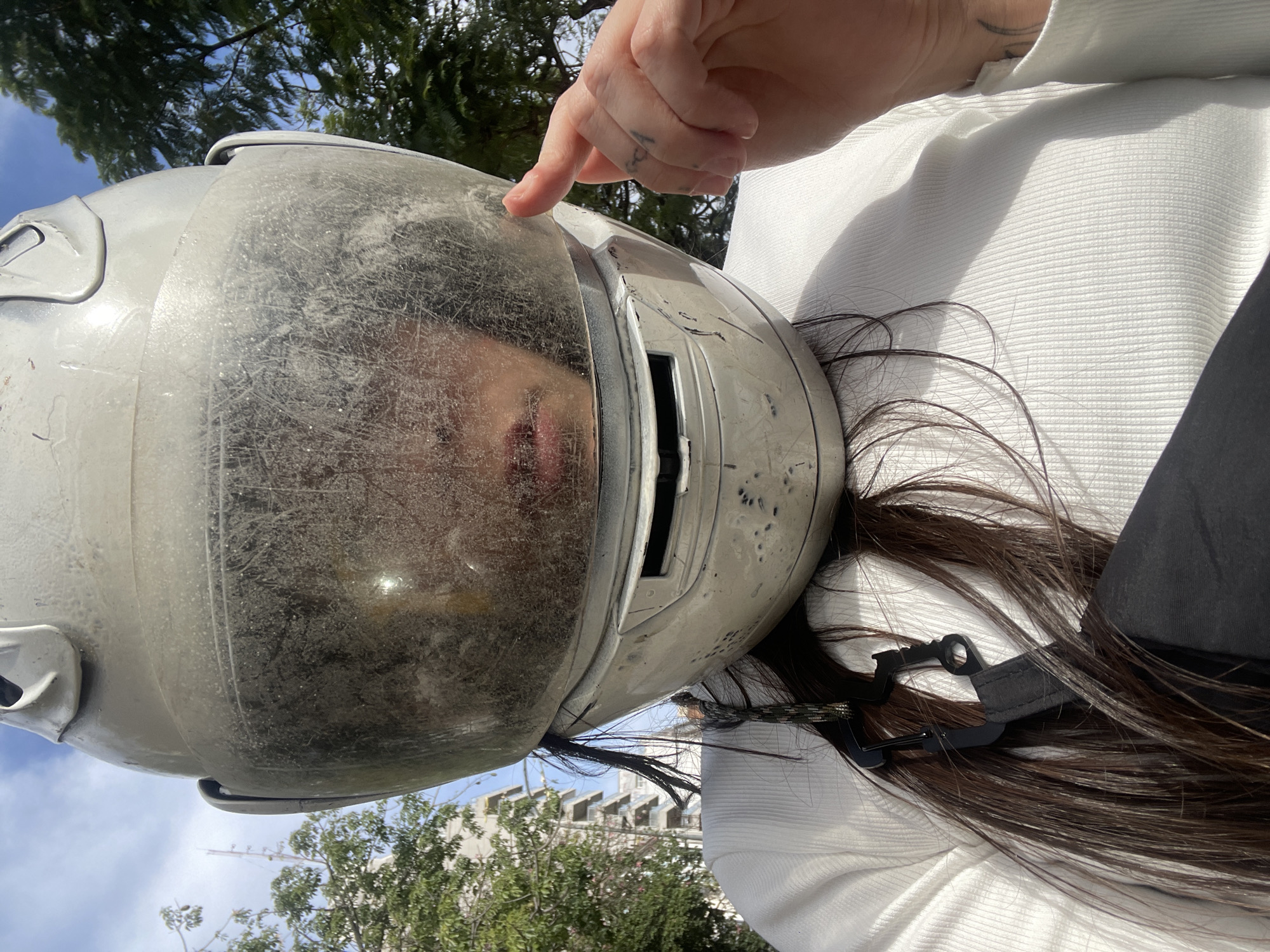 A person wearing a silver full-face helmet is seen from a close, angled perspective, with a finger pointing to the helmet. The helmet's visor is dirty and shows the reflection of the person's face. The person is wearing a white long-sleeved shirt. The background includes greenery and a building. Based on the architectural style, building height, and greenery, the location is possibly a neighborhood within Buenos Aires, Argentina.