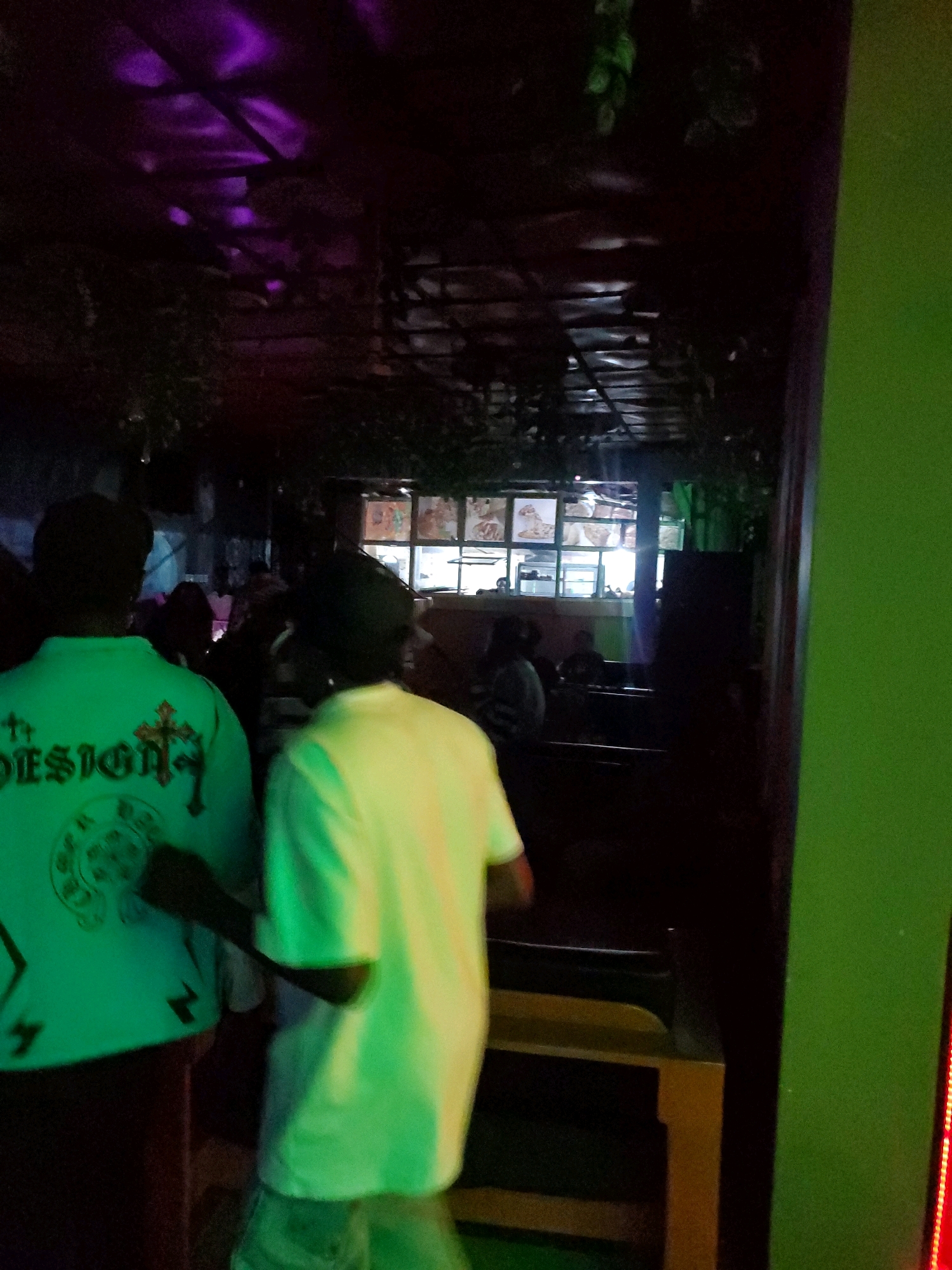 The image depicts an indoor scene at night, possibly a bar or restaurant, with purple lighting illuminating the ceiling and green lighting on the right side of the frame. Two individuals are visible from the back, facing away from the camera. The person on the left is wearing a luminous green long-sleeved shirt with a cross and the word "DESIGN" printed on the back. The person on the right is wearing a light yellow long-sleeved shirt. In the background, there are windows with what appear to be food displays or menus. The overall atmosphere is dimly lit and appears to be a social gathering place. The location is identified as the city of Jos, Nigeria.