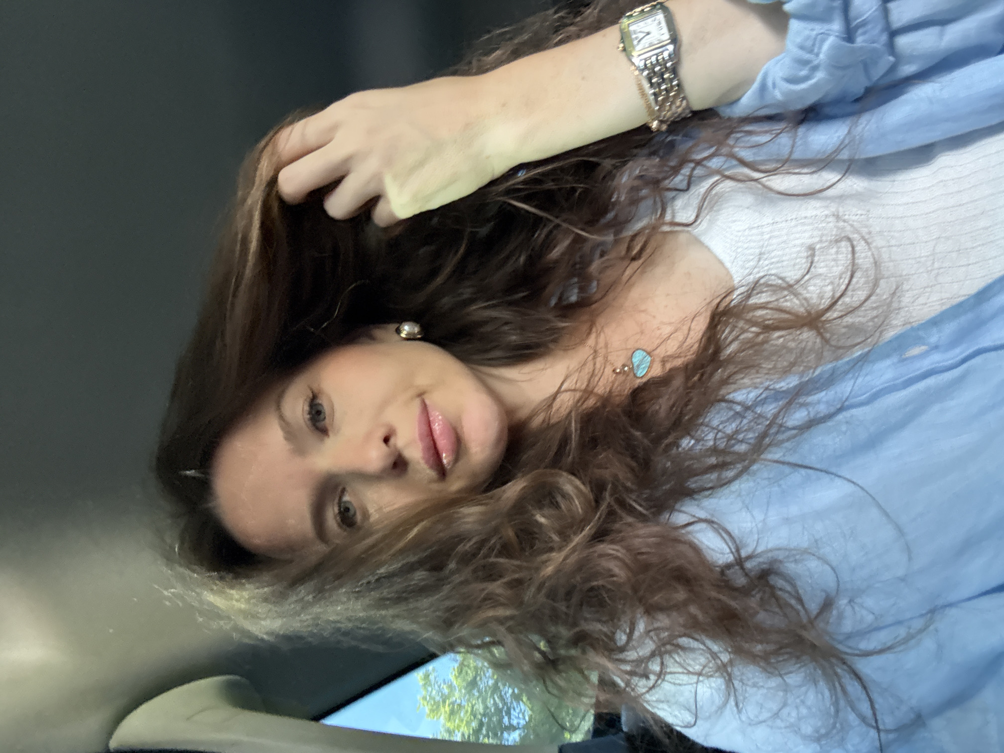 The image, oriented ninety degrees clockwise, captures a woman with long, wavy brown hair, likely posing for a selfie from within a car, given the provided location context of Moscow, Russia. Her head is tilted, and she looks directly at the camera with light-colored eyes and slightly parted, glossy lips.
She wears a light blue top over a white inner garment. On her left wrist is a gold-toned wristwatch with a rectangular face and visible numbers/markings, though no specific brand text is legible. She also has a small, pearl-like earring in her right ear and a necklace with a light blue, possibly turquoise, heart-shaped or irregular pendant. Her right hand is raised, with fingers gently touching her hair near her temple.
The background above her head is dark grey, resembling a car ceiling, while below her, through what appears to be a car window, green trees and a bright blue sky are visible, indicating it is daytime. Parts of the car's light-colored interior, possibly a handle or headliner, are also visible in the lower left. The overall mood is calm and natural.