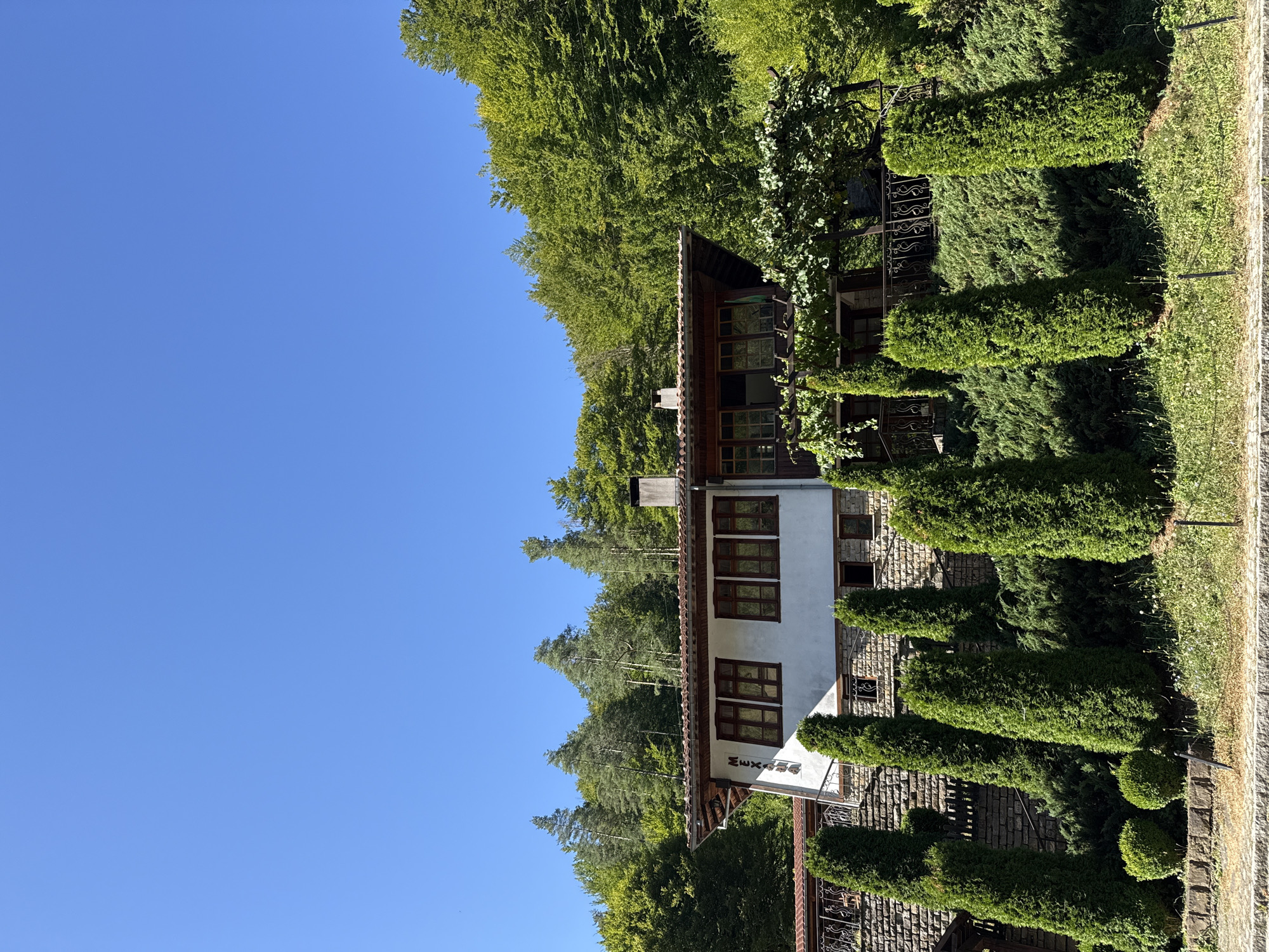 The image depicts a white, two-story building with a terracotta-tiled roof, nestled into a hillside. The building has numerous wooden-framed windows and a prominent stone foundation. It is surrounded by lush green trees and manicured cypress trees and bushes. A stone wall supports the lower level of the building, and a wrought-iron railing is visible. The sky is a clear, vibrant blue. The building is identified by the word "MEXANA" in large letters on its facade. This building is the Mechana Zelenika restaurant, located in the Etara Architectural-Ethnographic Complex near Gabrovo, Bulgaria.