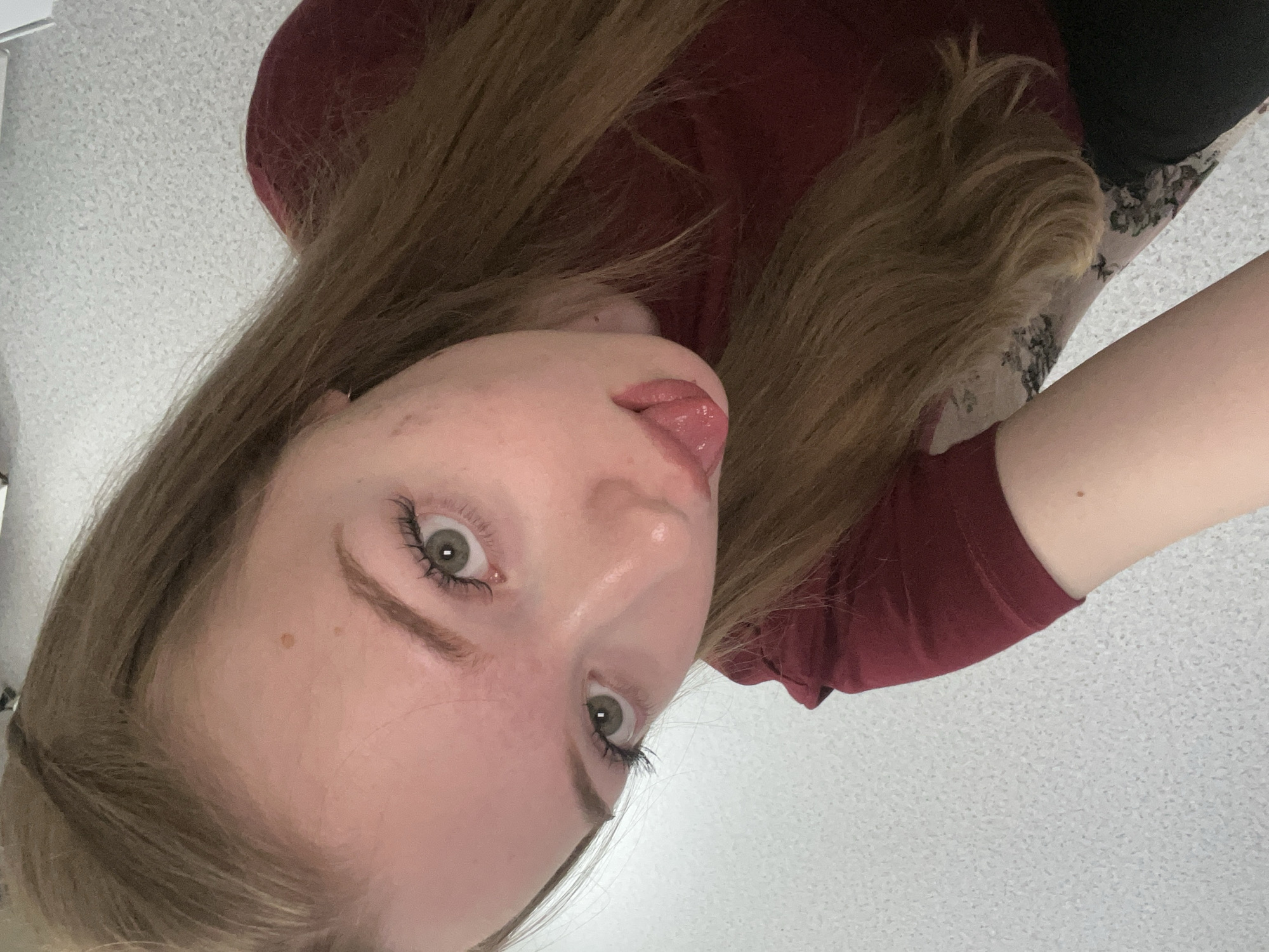 The image shows a young woman with light brown hair and green eyes. She is wearing a maroon-colored shirt. The background is a light gray speckled surface, which might be a wall or countertop. The woman is sticking her tongue out slightly. The photo appears to be taken at a close-up, angled perspective. The location is not identifiable as Chaikovsky, Russia based on the image alone.