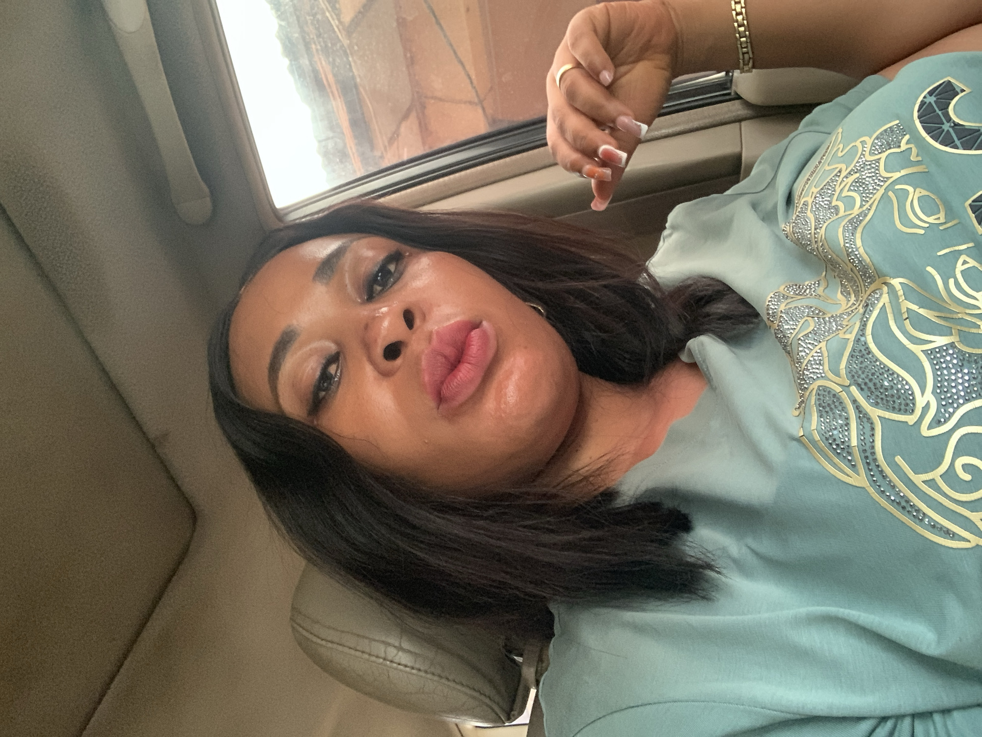 A woman with dark hair and a light green top is positioned inside a vehicle, likely a car. The location is speculated to be Sabon Tasha, Nigeria. The woman has a gold ring on her finger and a gold bracelet on her wrist. She is looking at the camera with her lips pursed. The interior of the car is beige. A window is visible in the background. The weather appears to be overcast outside.