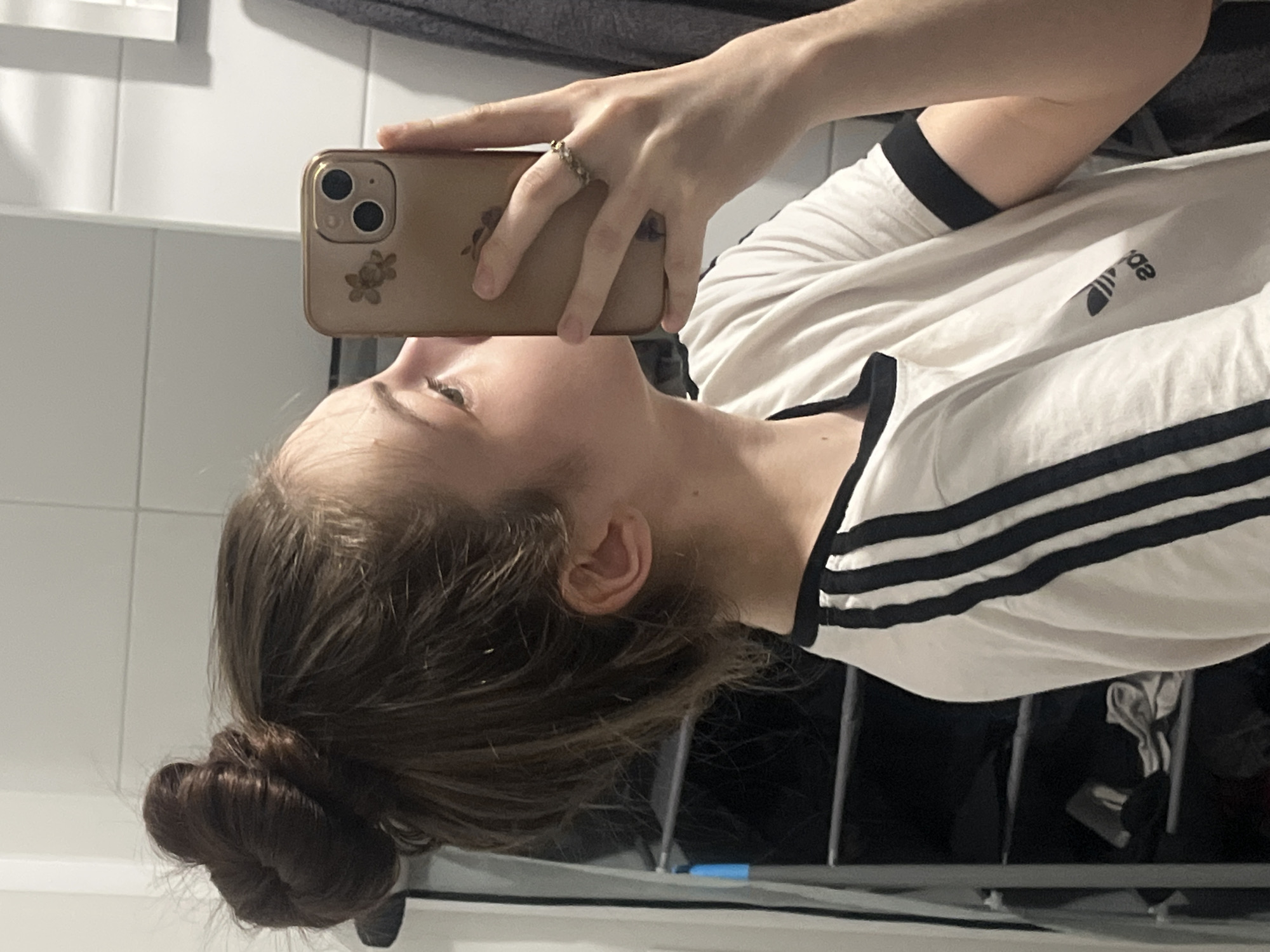 A person is seen from the left profile, capturing a mirror selfie indoors. The individual has light skin and dark brown hair styled in a high bun. They are wearing a white t-shirt with black trim on the collar and sleeves, featuring three black stripes along the shoulder and a partially visible, reversed Adidas trefoil logo on the chest area. A dark undershirt is visible beneath the t-shirt neckline. The person holds a gold-colored smartphone with a floral-decorated case and a triple camera setup in their left hand, which displays a patterned ring on the ring finger and a plain band on the middle finger. The reflected background shows a white tiled wall, a dark gray towel hanging on the far right, and a dark shelving unit on the left with various folded items and a blue object.