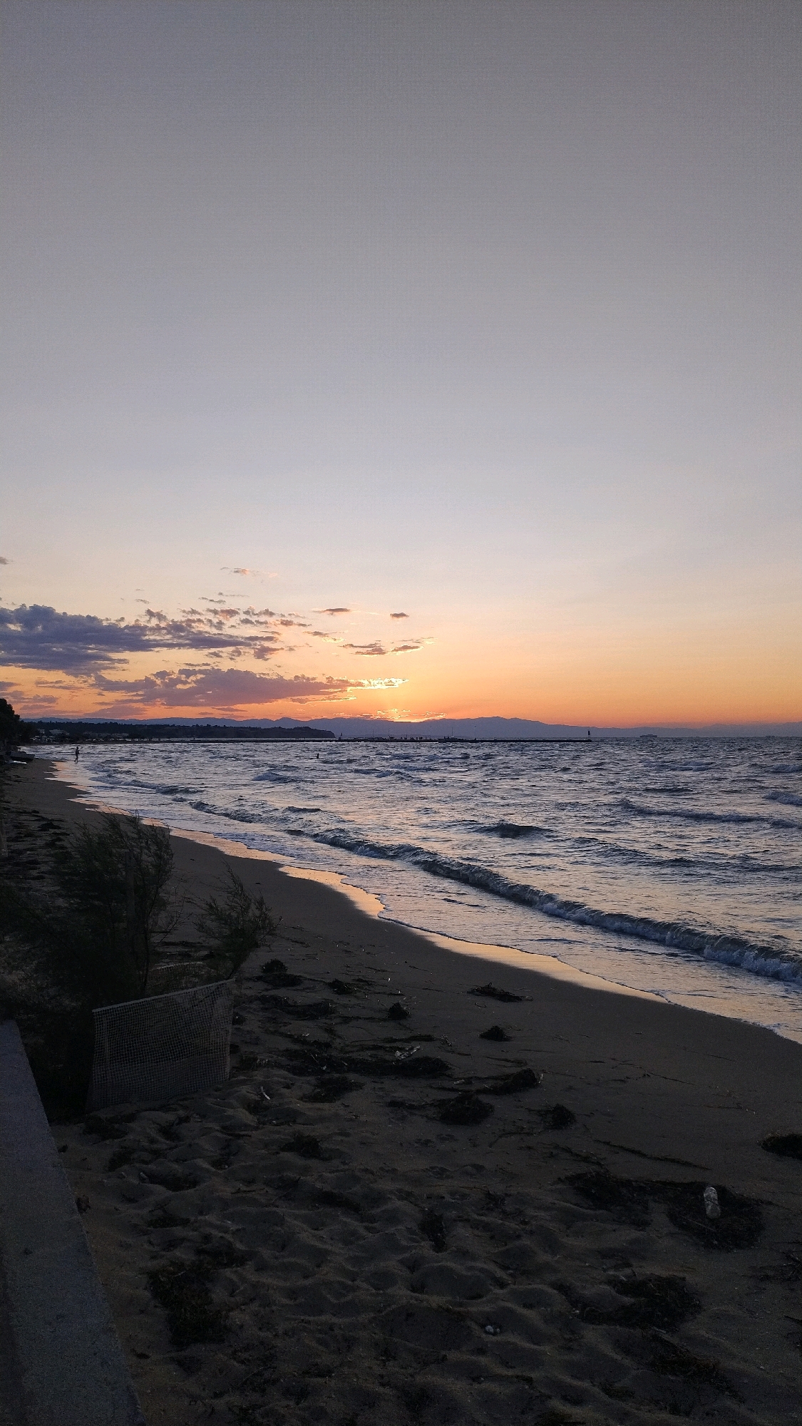 The image depicts a sunset scene at a beach in Neoi Epivates, Greece. The sky is a gradient of soft purples and oranges, with the sun partially visible just above the horizon, casting a warm glow on the clouds and water. The sea is calm with gentle waves lapping at the sandy shore. The beach itself is dark, with scattered debris and vegetation. In the distance, a line of mountains can be seen against the twilight sky, and a pier or jetty extends into the water. There are a few people visible on the beach and in the water, appearing as small silhouettes against the bright sky. A mesh bin is also visible on the sand.