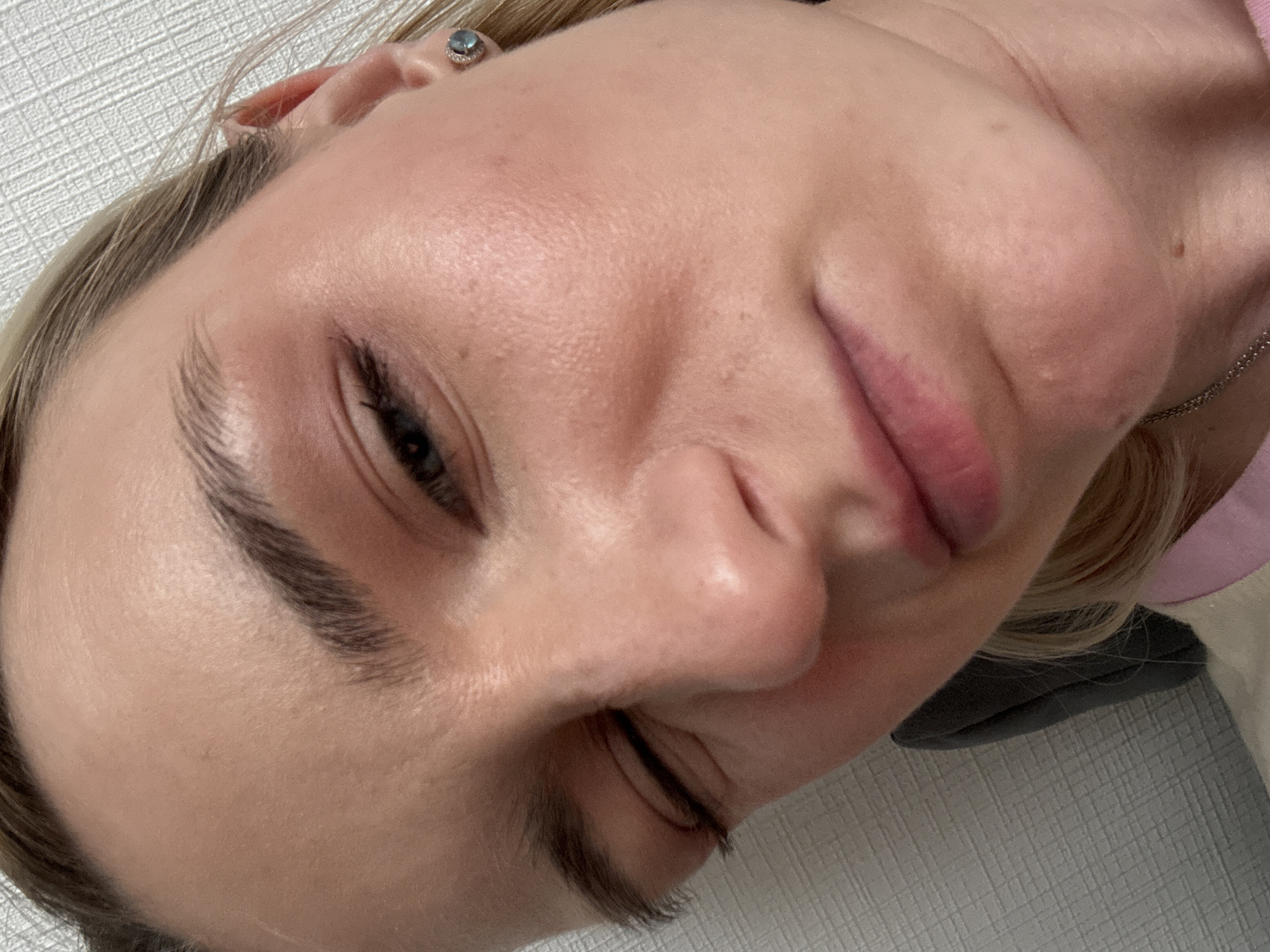 A close-up, angled headshot of a fair-skinned person with blonde hair. The person is angled with their right side slightly higher. Their right eye is open and shows dark eyelashes and a blue-grey iris. The right eyebrow is dark and thick. The nose is prominent. The lips are full and pink. A silver earring with a blue stone is visible in their right ear. A thin silver chain is visible on the right side of their neck. The background shows a white textured surface. The location in the city of Vsevolozhsk, Russia is not detectable from this image.