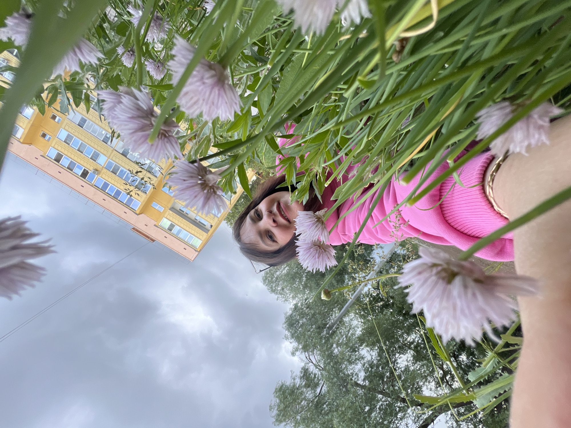 This image captures a close-up, upward-angled perspective of a young woman smiling through a bed of light purple flowers and green foliage. The woman, with dark hair and glasses, wears a bright pink ribbed long-sleeved top and a gold-colored bracelet on her right wrist. She is looking directly at the camera with a happy, content expression, her face partially framed by the abundant blossoms and leaves in the foreground.
Behind the person and the greenery, a large, multi-story yellow building with numerous rectangular windows fills the left side of the frame. The architecture suggests an apartment block or similar urban structure. Green trees are visible in the mid-ground below the building. The sky above is overcast with light gray clouds, indicating a cloudy daytime setting. The camera appears to be positioned very low to the ground, looking up through the plants, giving a unique, immersive view. No visible text is present in the image. The scene is set in Noviy, Russia, showcasing a blend of urban environment and natural elements.