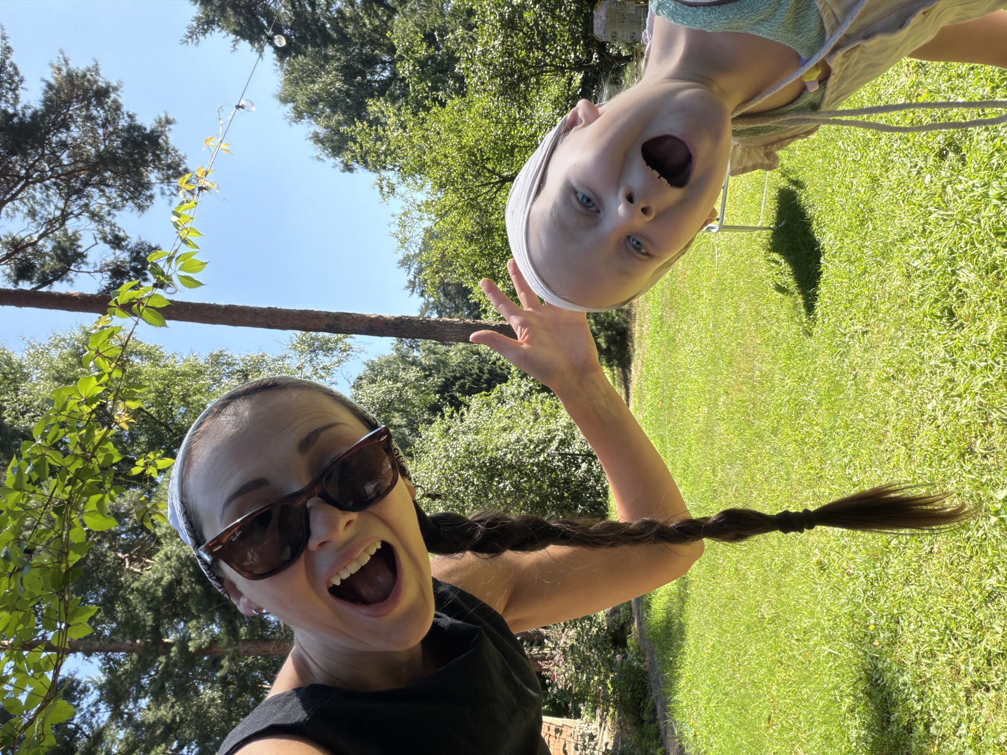 This media file features a selfie taken outdoors, likely in Udelnaya, Russia, given the provided context. Two subjects are visible: a woman in the foreground and a child in the background, both appearing to be taking the photo together. The woman is wearing sunglasses and a black top, with a white bandana on her head and her long, dark hair braided and extended outwards. She has an open-mouthed expression, conveying excitement or surprise. The child, positioned upside down relative to the woman, is also looking at the camera with a wide, open-mouthed expression, wearing a light-colored garment and a white bandana. The setting is a sunny outdoor environment with lush green grass and trees, suggesting it's daytime with pleasant weather. A thin wooden pole and some hanging string lights are visible in the upper left portion of the frame, adding to the outdoor ambiance. The overall mood is playful and joyful, capturing a moment of shared fun between the two individuals.