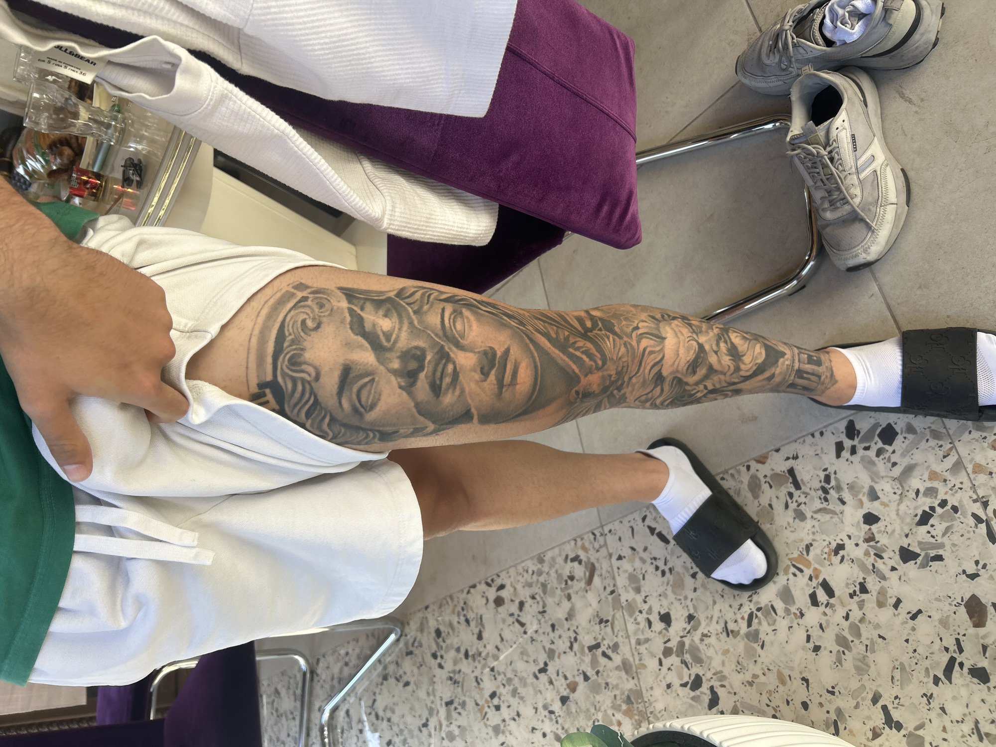 A man is seen from an overhead perspective, wearing white athletic shorts, a green shirt, white socks, and black Gucci slides. His right leg is heavily tattooed with a predominantly black and gray design featuring faces and ornamental patterns. The setting appears to be indoors on a light-colored speckled floor. Several items are scattered around, including a pair of gray sneakers and a stack of white towels or clothing on a purple cushion. Additionally, bottles with various contents are visible in the upper left corner. There is no information that points to the location of Elhovo, Bulgaria.