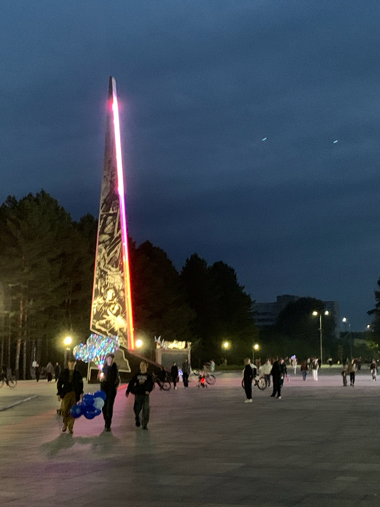 In Prokopyevsk, Russia, a monument illuminated with pink and red light stands tall against a twilight sky. The monument is adorned with intricate carvings. People are visible in the foreground, some walking and others standing. A group of three individuals in the lower left of the frame are carrying a bunch of blue and white balloons. The area appears to be an open plaza, with trees and streetlights visible in the background. Several bicycles and scooters are also present, suggesting a public space that is used for recreation. The overall atmosphere is that of an evening gathering in a city square.