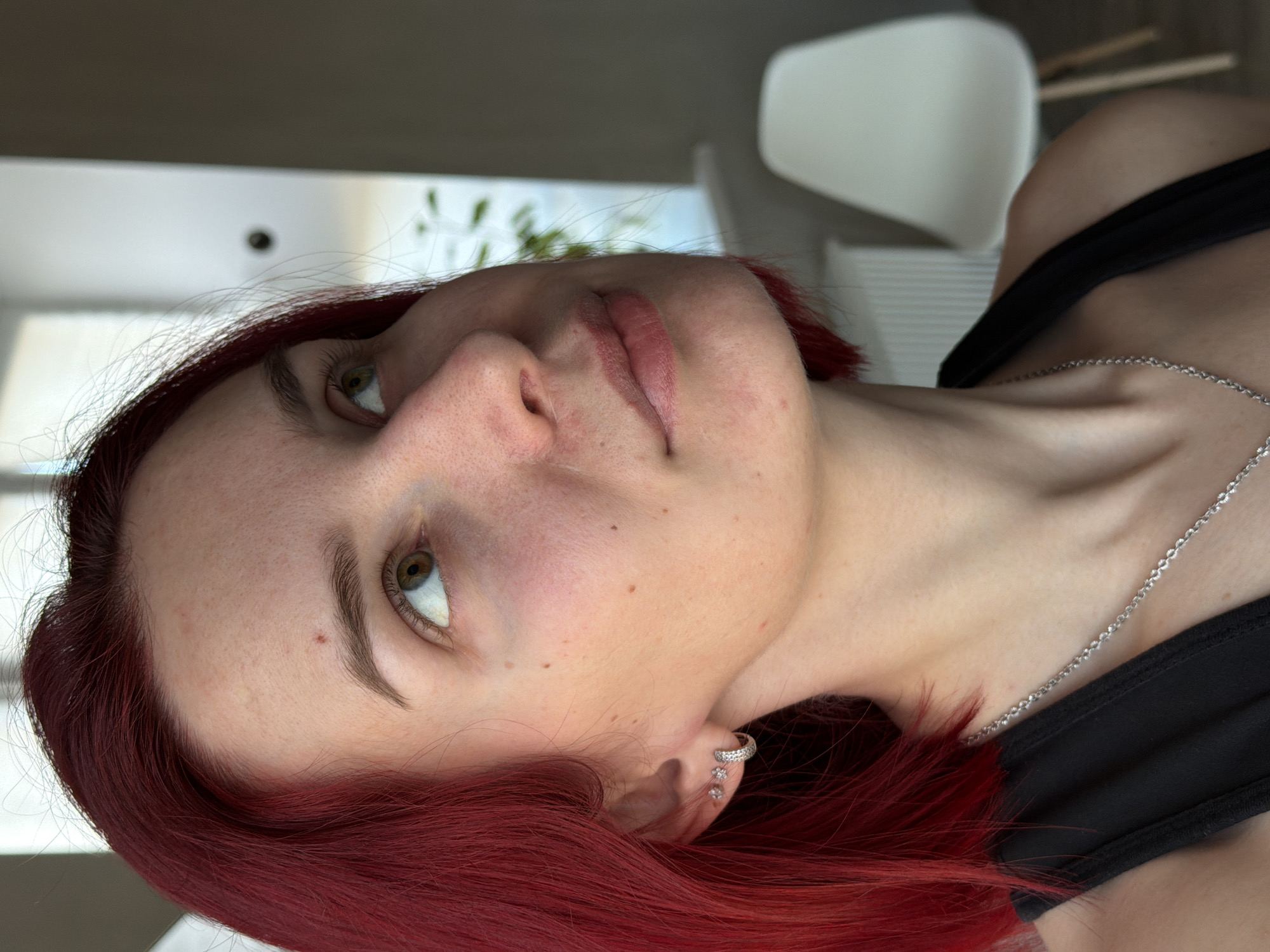 A close-up, angled portrait of a fair-skinned woman with red hair. She wears a black top and a silver necklace. Her left ear has a sparkling earring. The background shows a blurred interior with a white chair and a window with greenery. There is no indication of the location of the city of Chaikovsky, Russia, in the image.