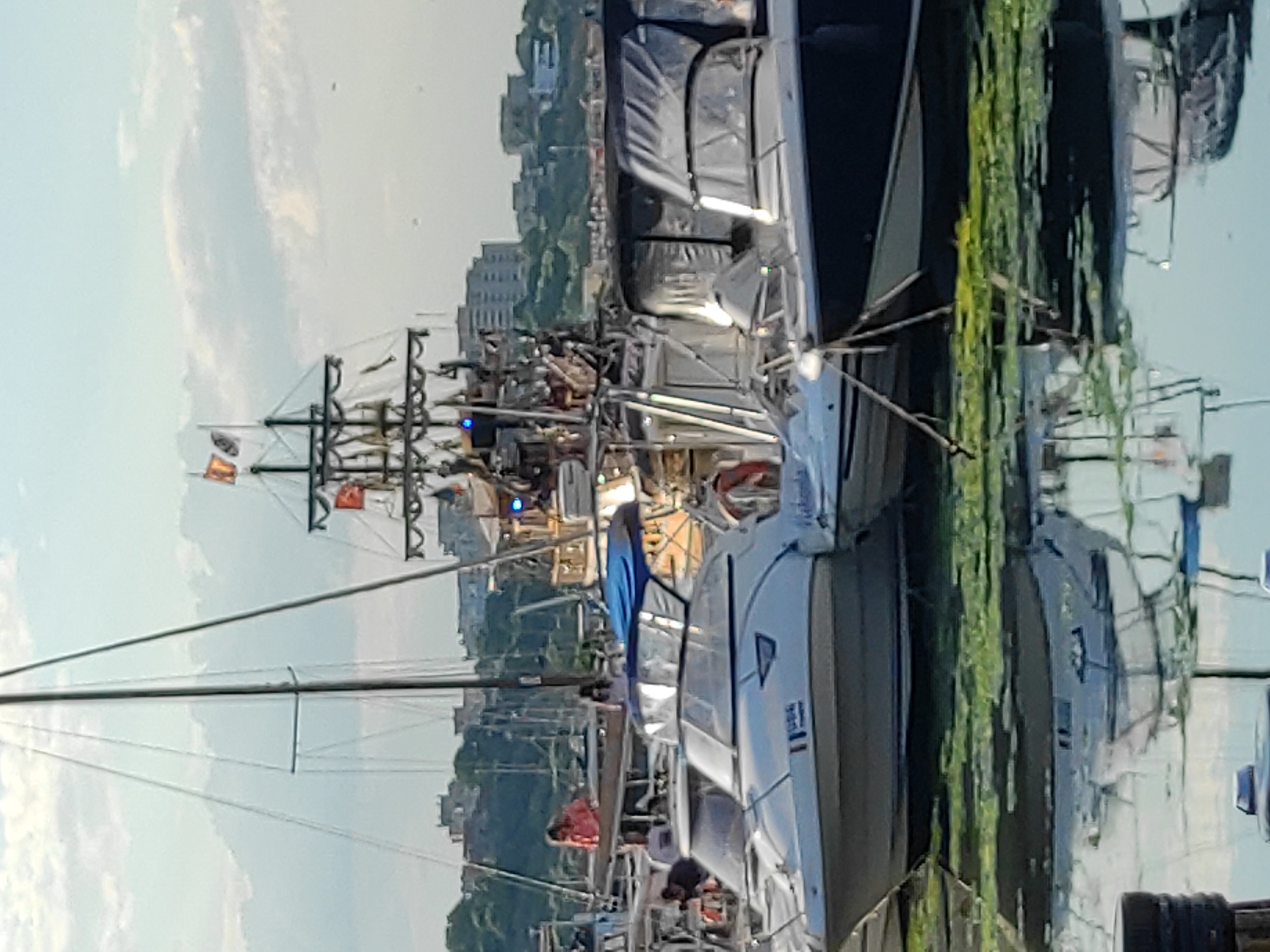 The image depicts a harbor with several boats docked, including a large, multi-decked vessel resembling a pirate ship adorned with flags and lights. Smaller pleasure boats with cabins are also visible, some with their canopies up. The water is partially covered with green algae or aquatic plants, and reflections of the boats and the sky are visible on the surface. In the background, buildings are seen on a hill overlooking the harbor, suggesting an urban setting. A person in a pink shirt is standing on one of the boats in the mid-ground. The sky is partly cloudy. The scene appears to be the Constanța Old Port in Constanța, Romania, known for its mix of historical and modern vessels.